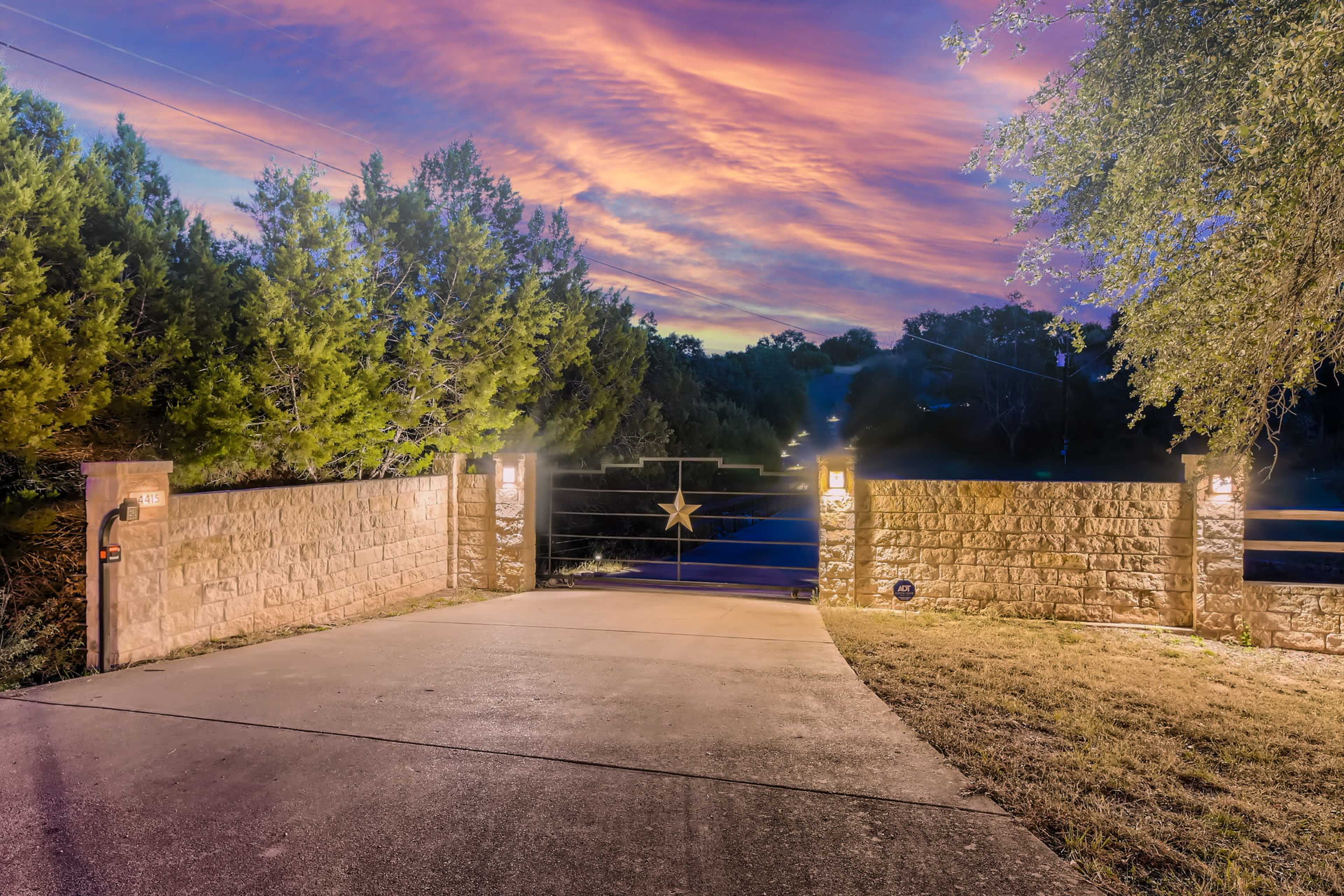 A stone entrance gate with a star design is illuminated by lamps, leading to a gravel driveway amidst trees under a vibrant sunset sky.