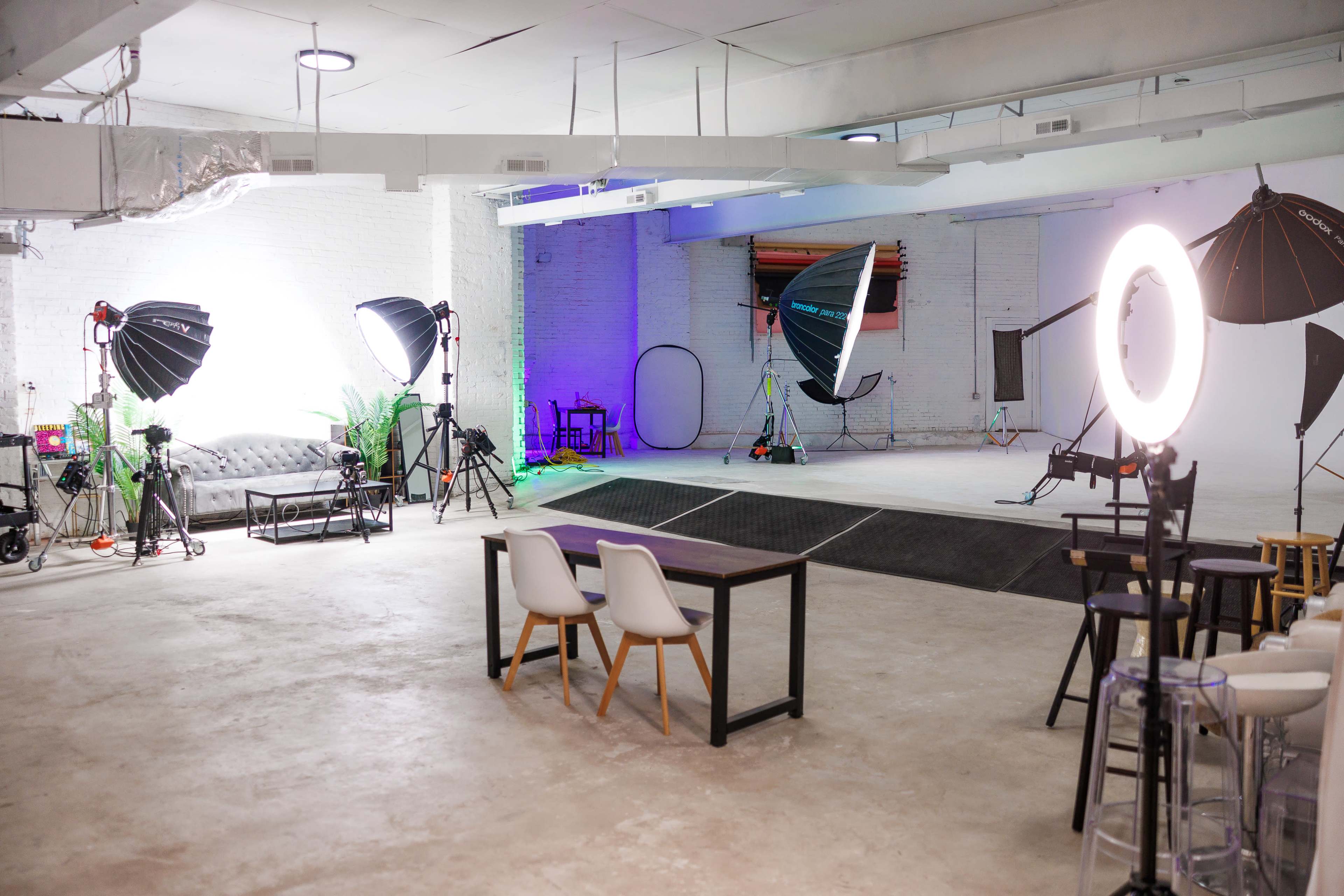 The image shows a spacious photography studio equipped with various lighting setups, a gray couch, a table, and a backdrop area.