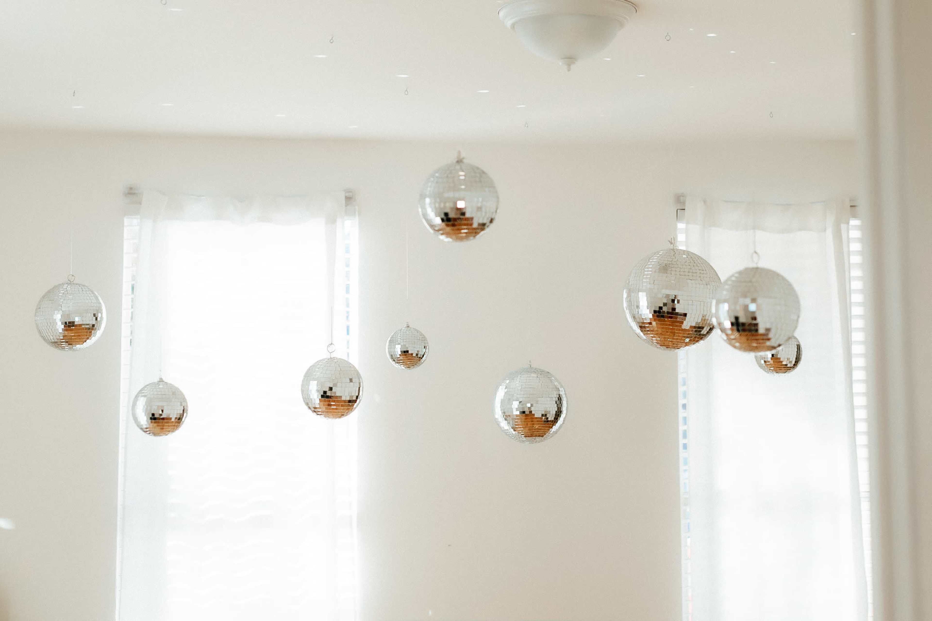 Several disco balls hanging from the ceiling in a room with white walls and sheer curtains.