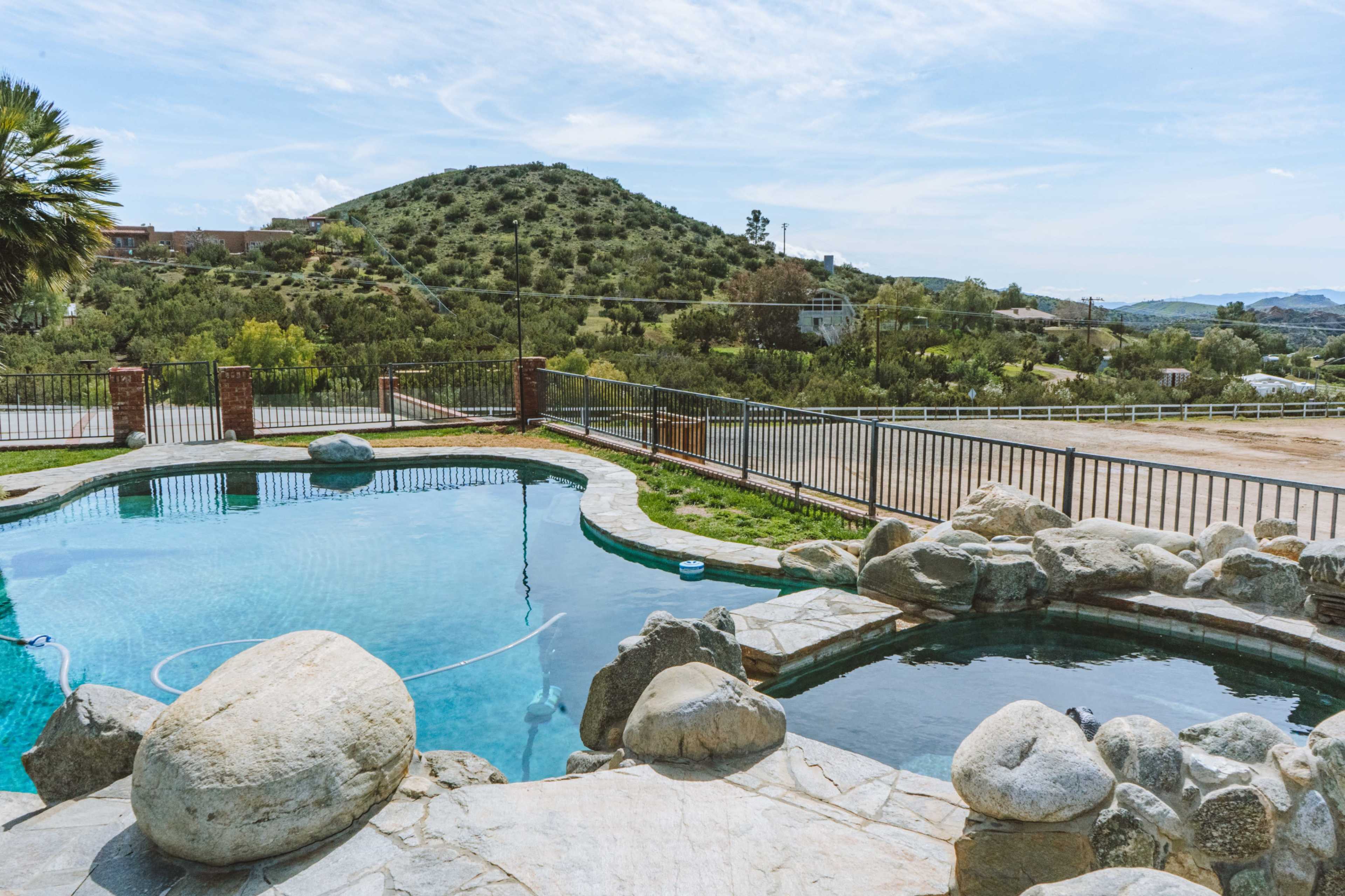 The image shows a swimming pool surrounded by rocks, with a hillside and a fence in the background.