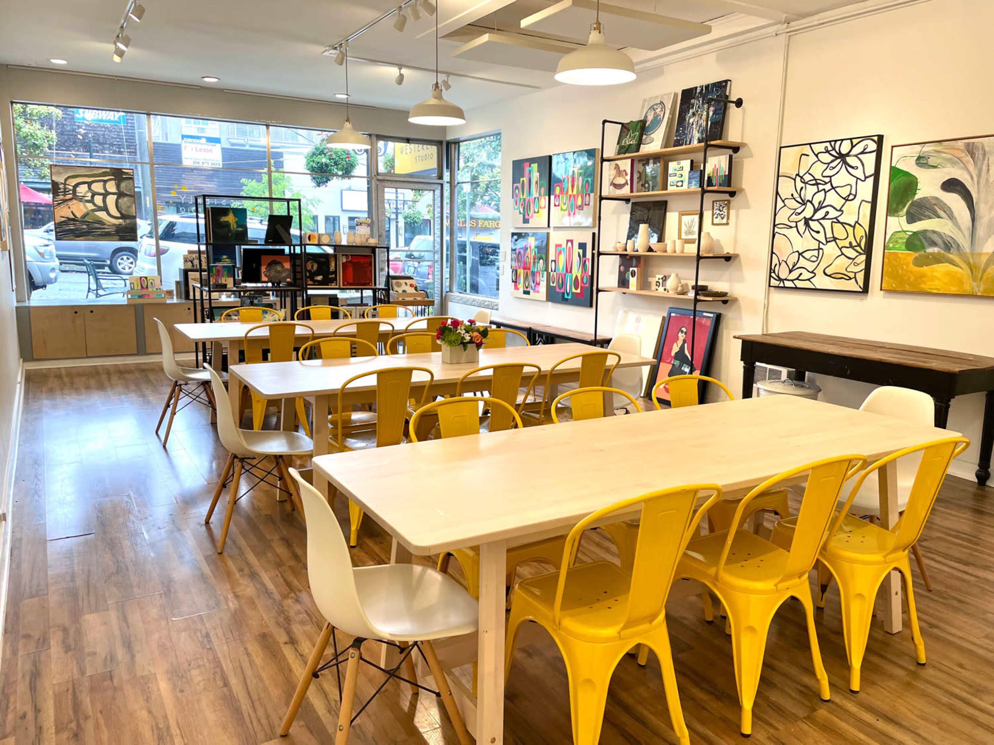 A bright art studio featuring multiple tables with yellow chairs, shelves displaying artwork, and large windows allowing natural light to enter.