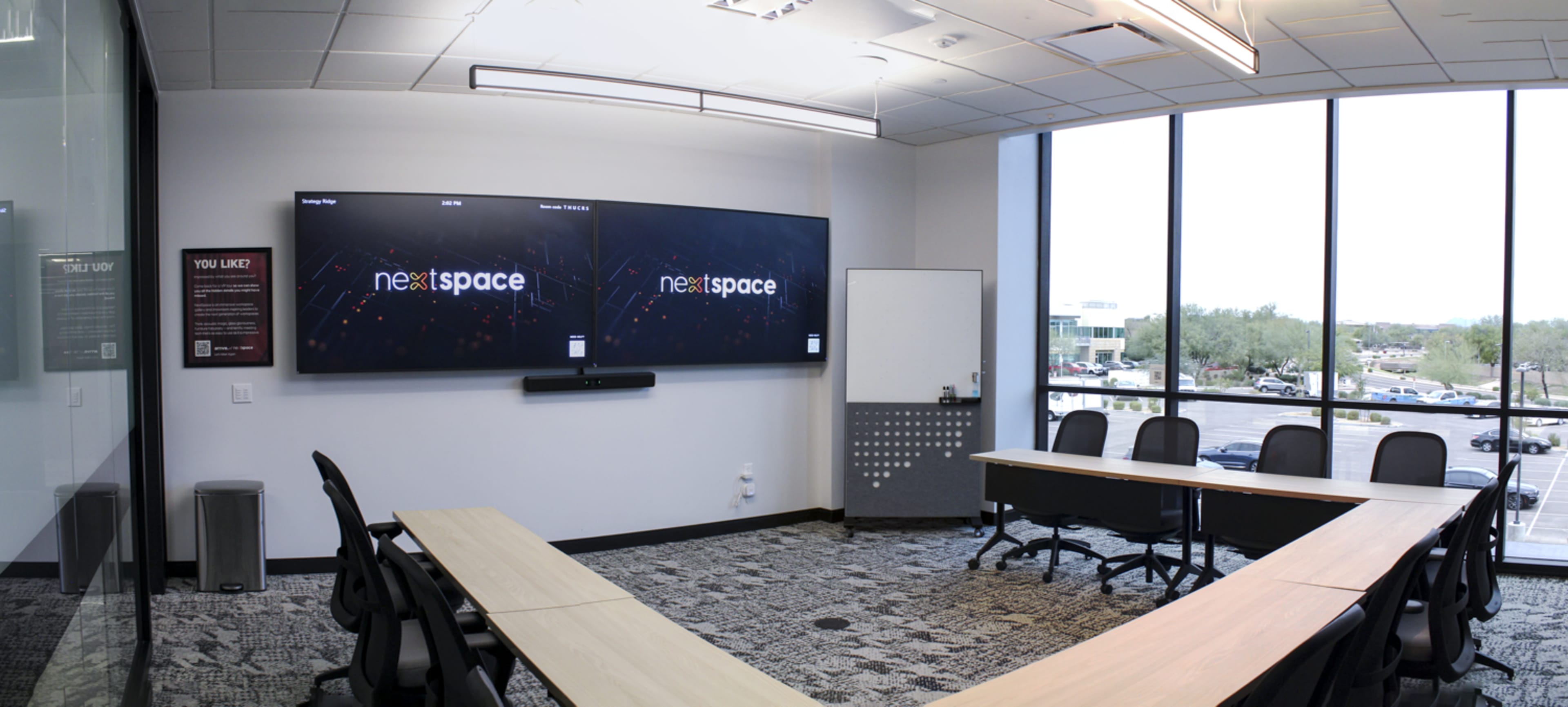A modern conference room features large screens on one wall, a U-shaped table, and large windows with a view of an outside parking area.