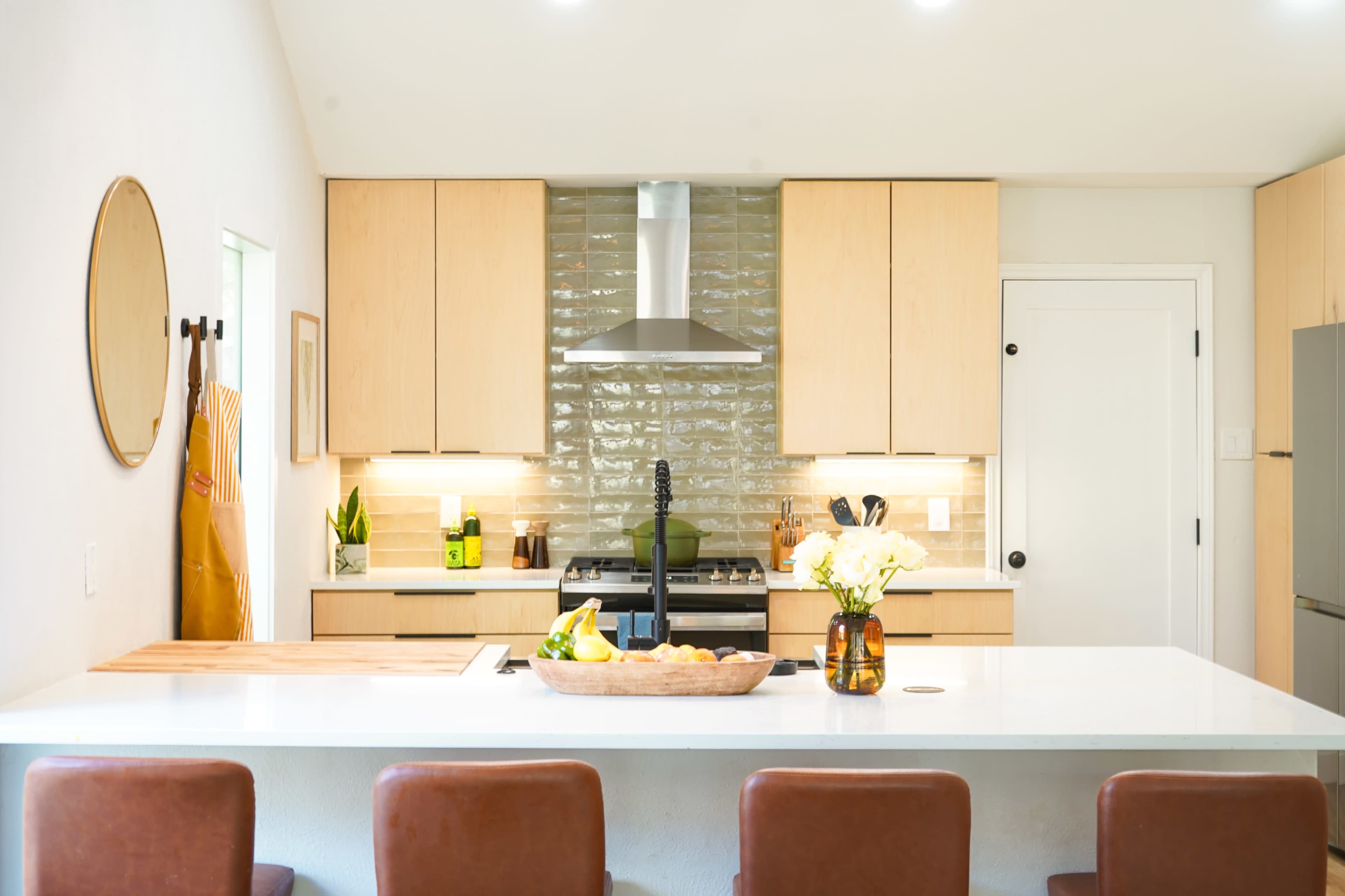 A modern kitchen features light wooden cabinets, a stainless steel range hood above a stove, and a central island with a bowl of fruit and flowers.