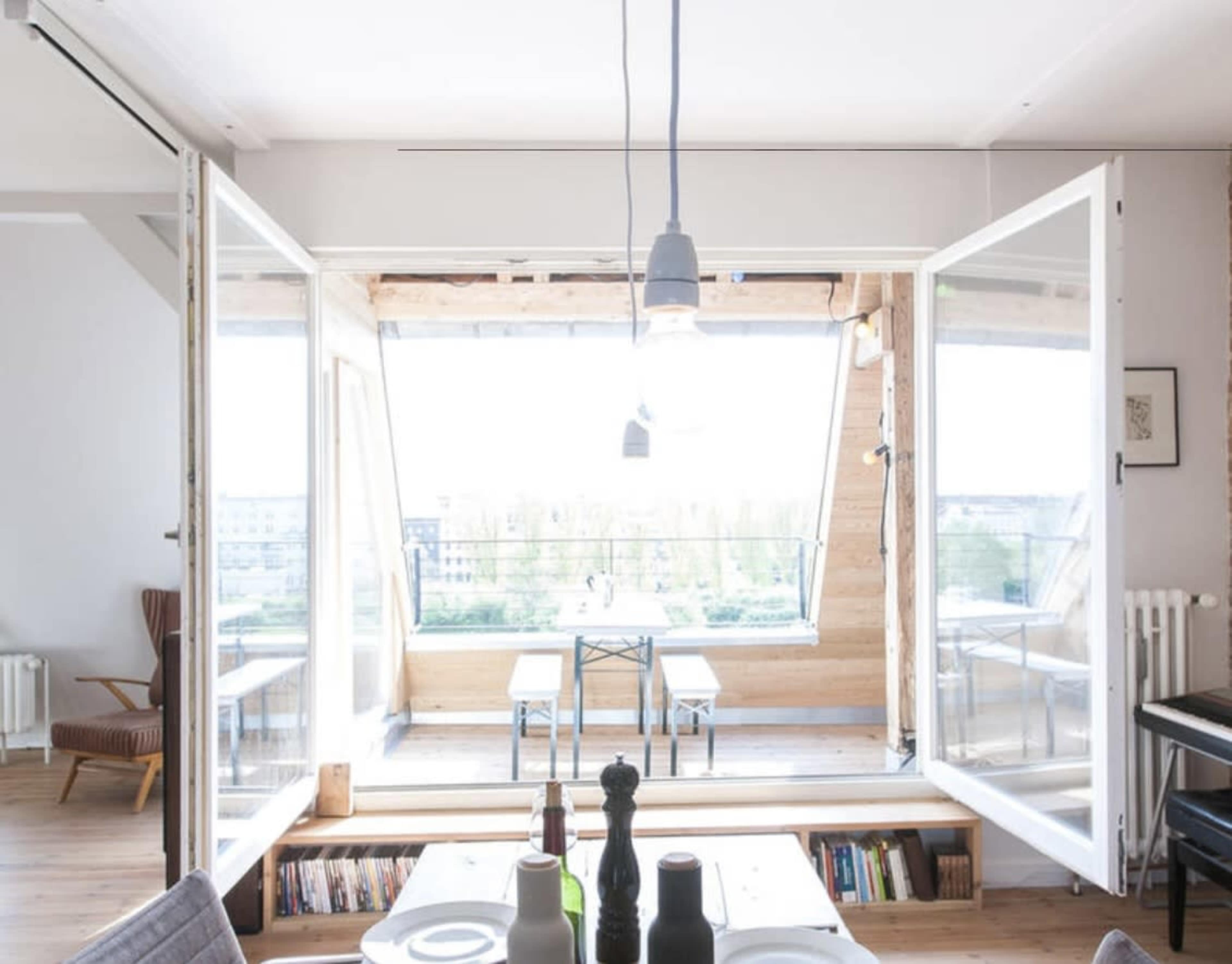A spacious room features large open windows that overlook a wooden terrace with a table and chairs, allowing natural light to flood the interior.