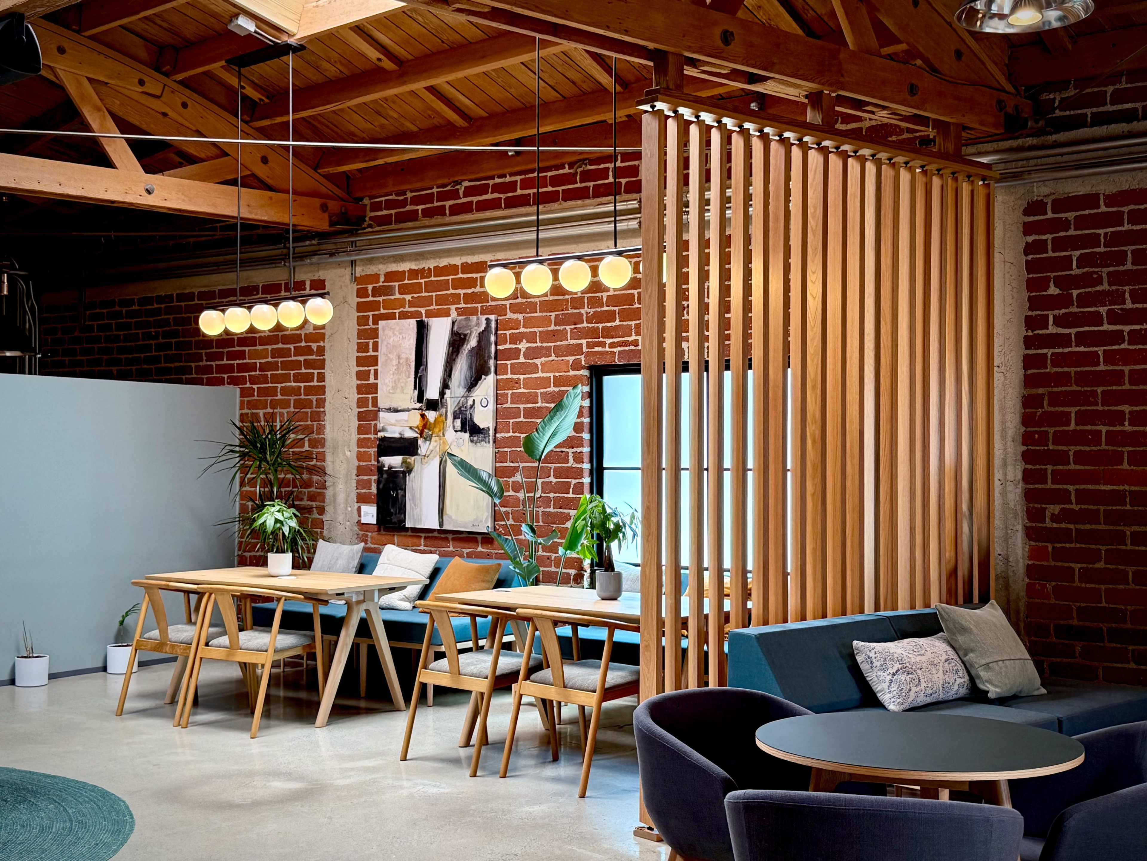 The image shows a modern workspace with wooden tables, a decorative wooden partition, and exposed brick walls, featuring plants and contemporary art.