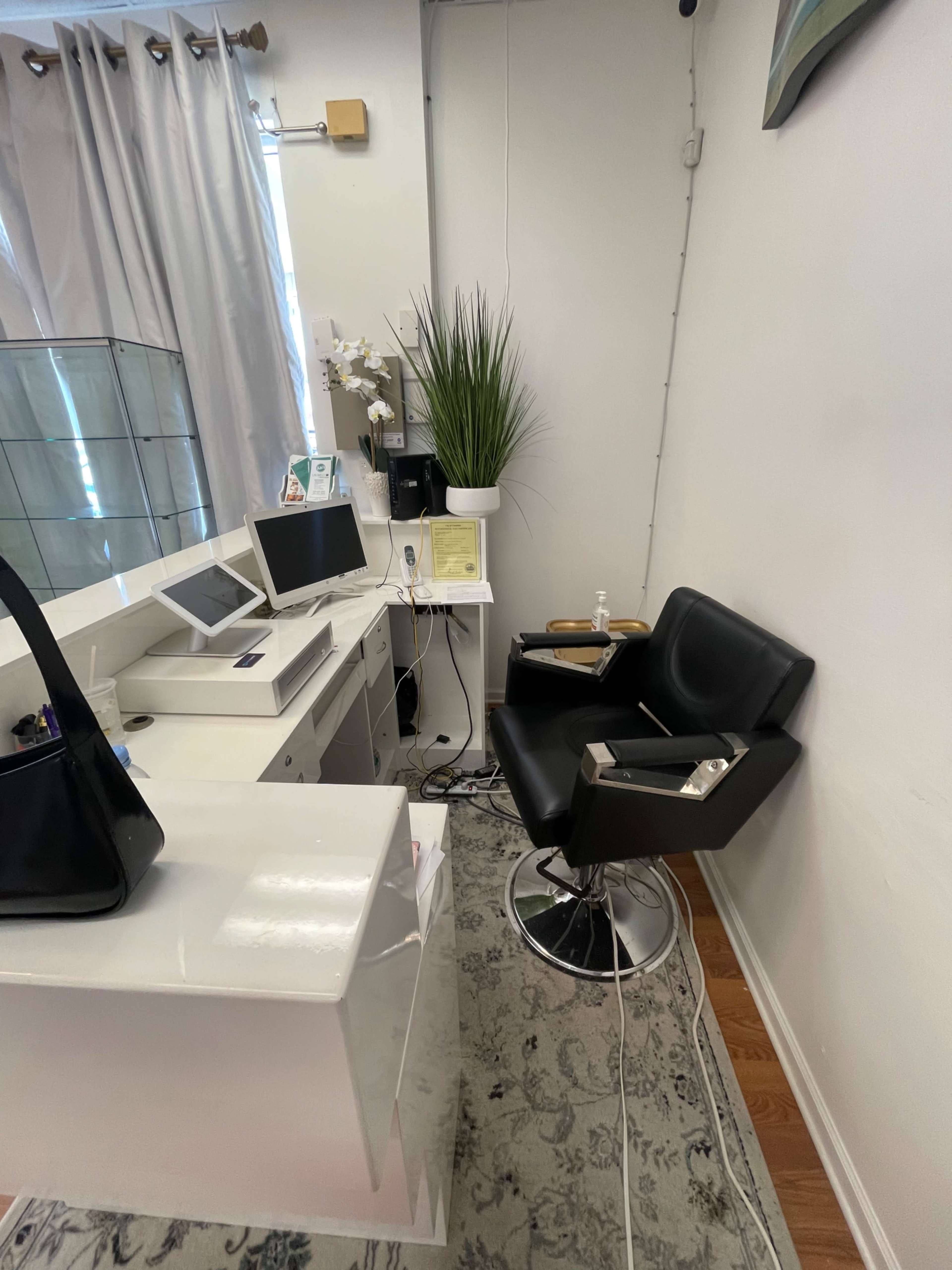 A small salon area featuring a black salon chair next to a white desk with electronic devices and decorative plants.