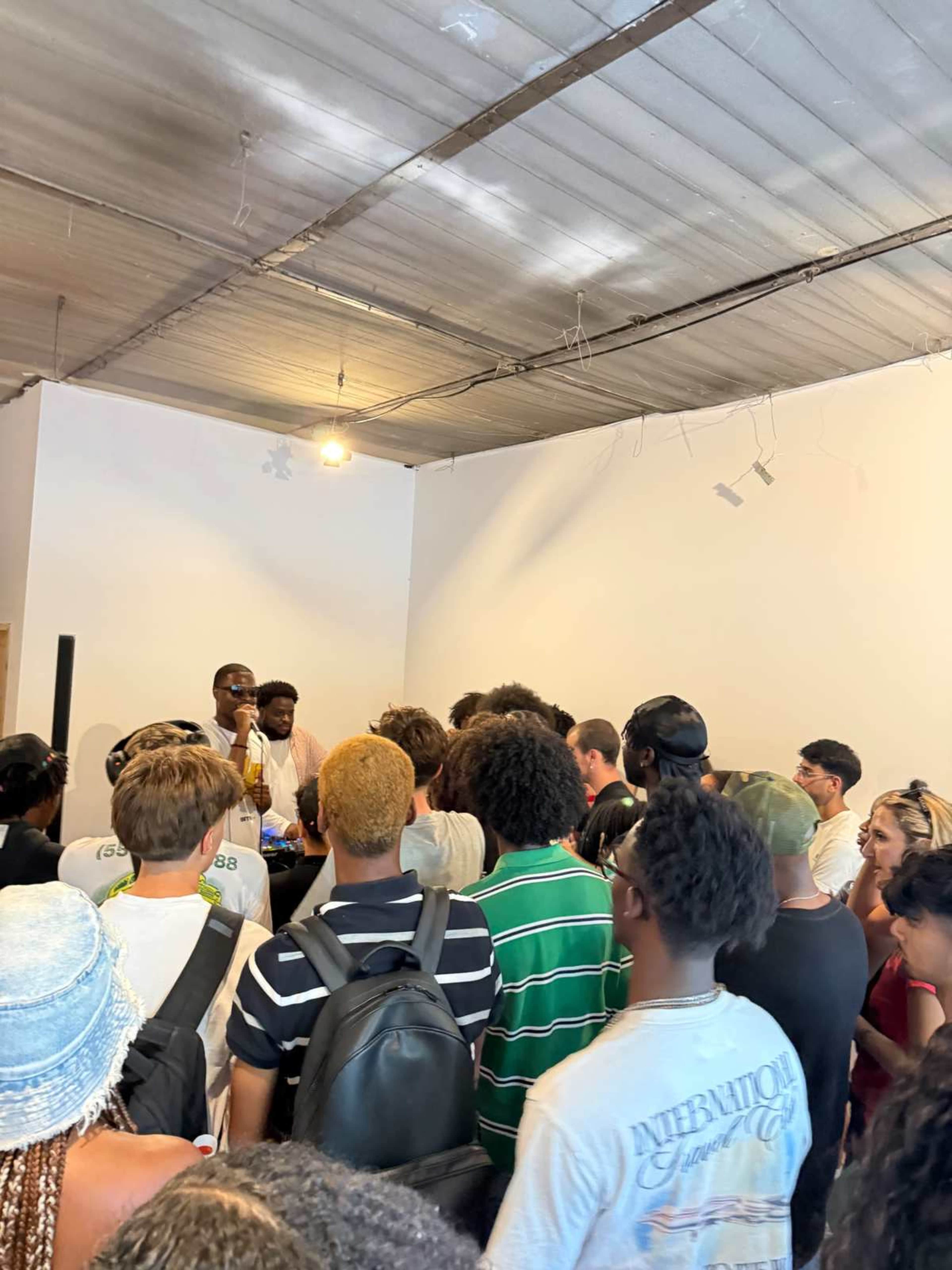 A crowd of people gathers around two speakers in a brightly lit room.