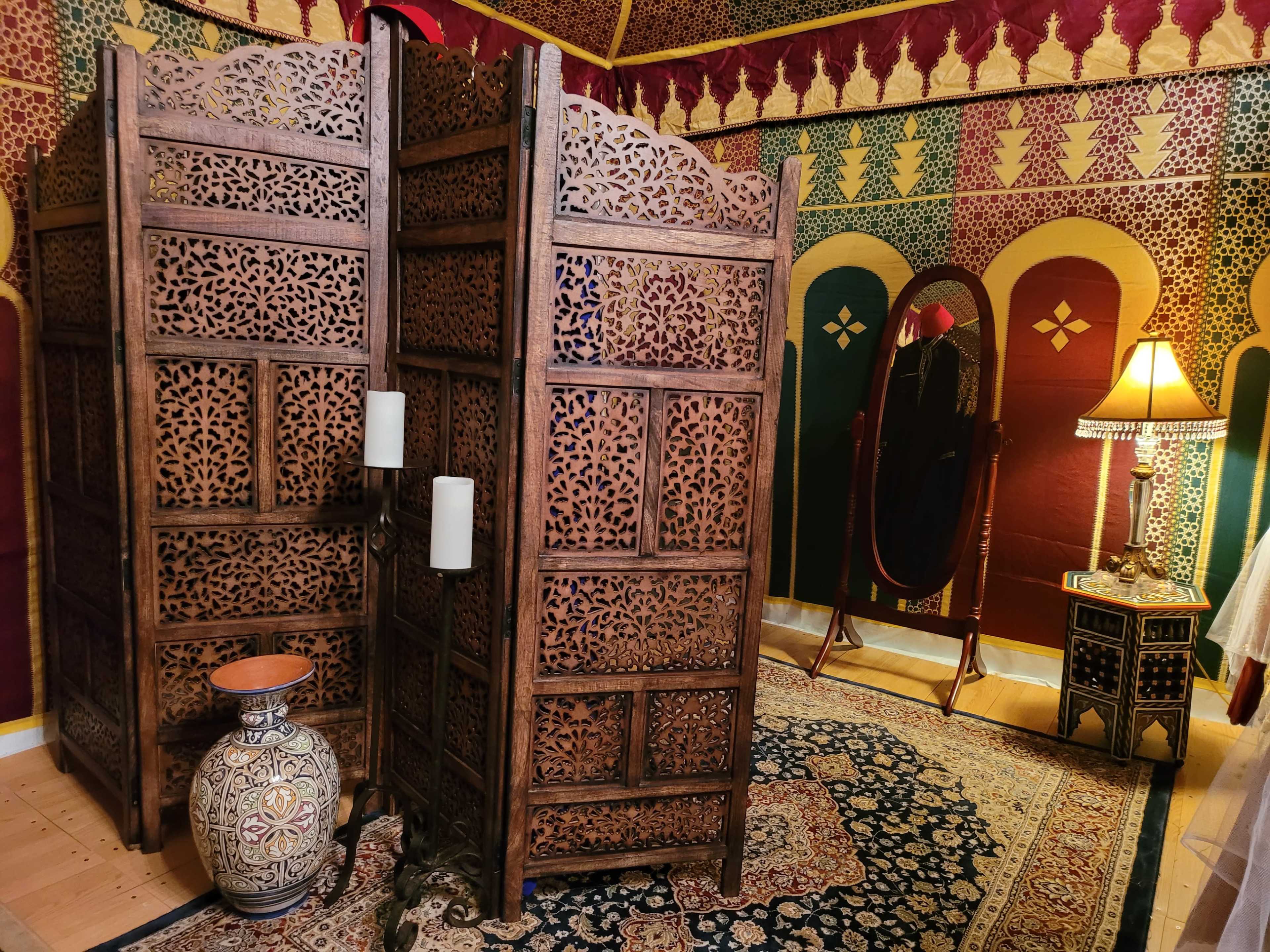 A decorative room with wooden screens, a standing mirror, a lamp, and an ornate vase on a colorful rug.