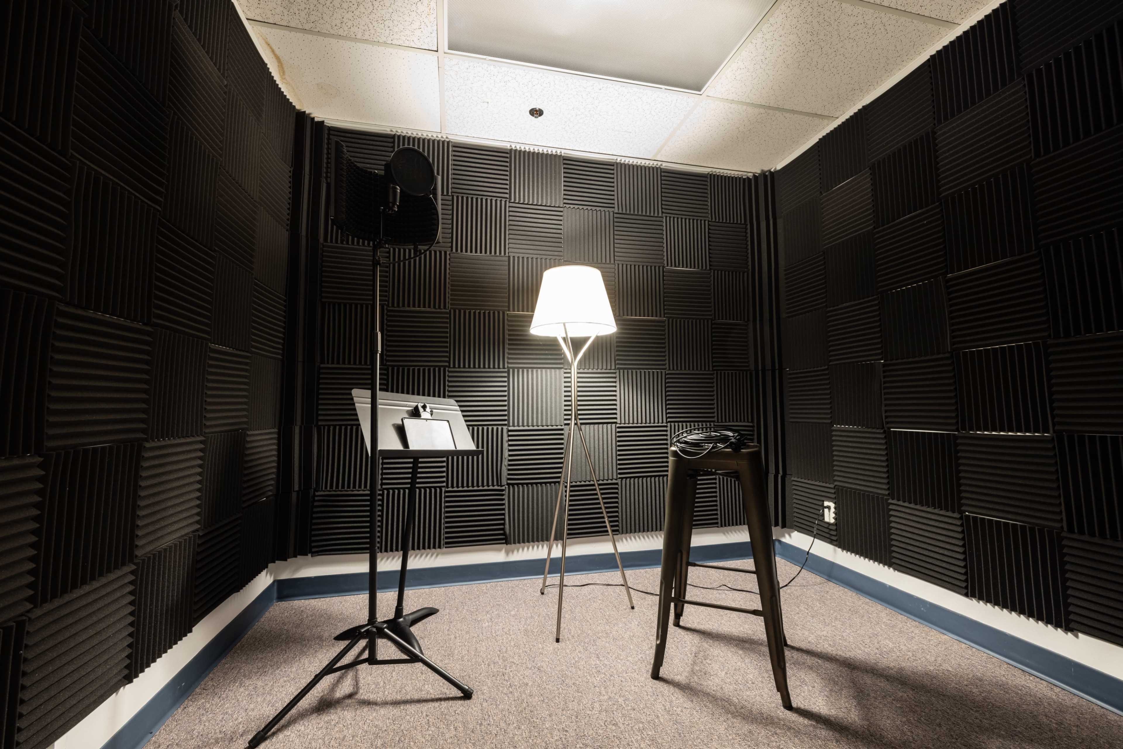 The image shows a soundproof recording studio with black foam acoustic panels on the walls, a microphone stand, a stool, and a floor lamp.