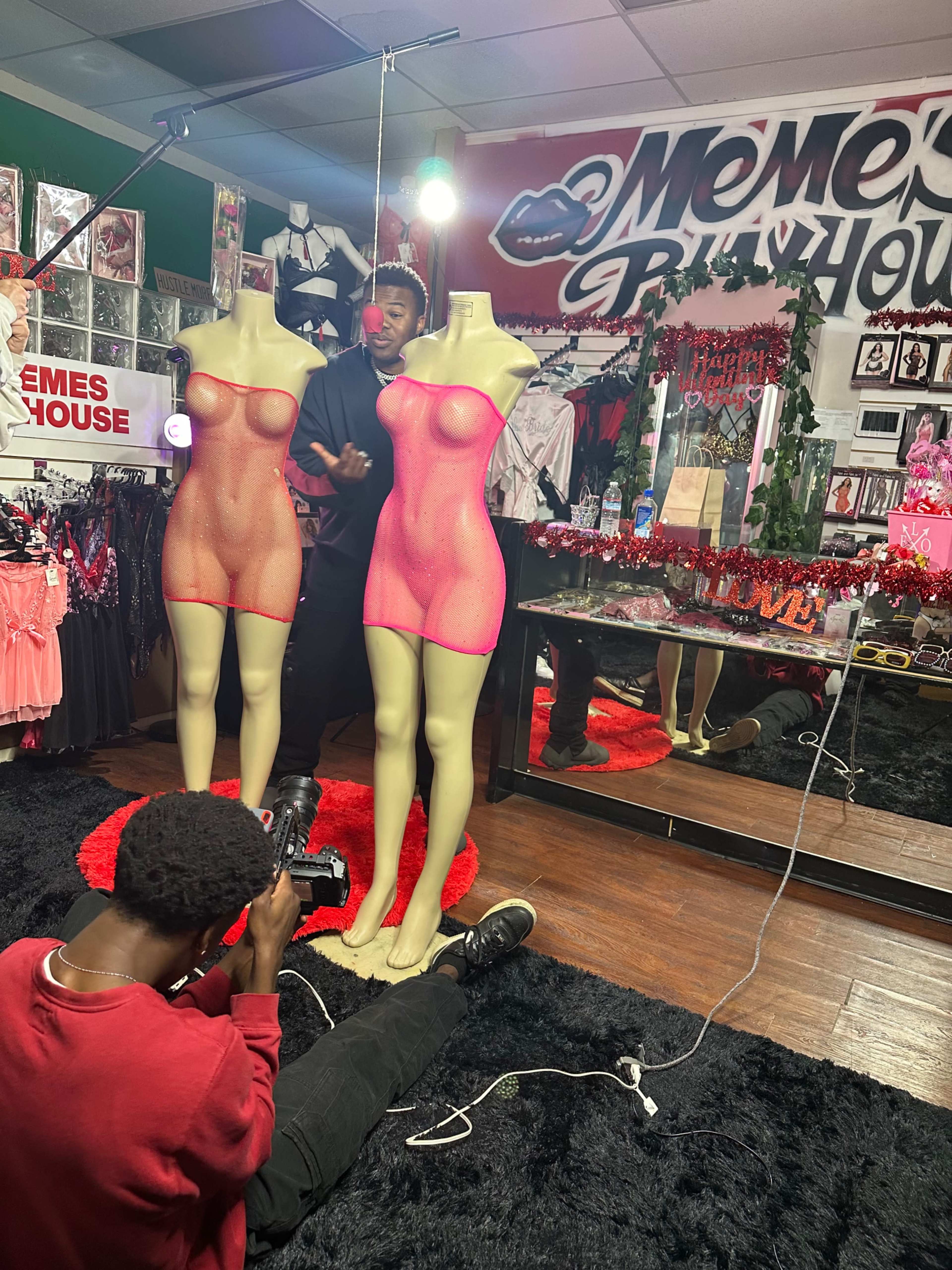A person sits on the floor photographing two mannequins dressed in sheer pink outfits in a store filled with colorful decorations.