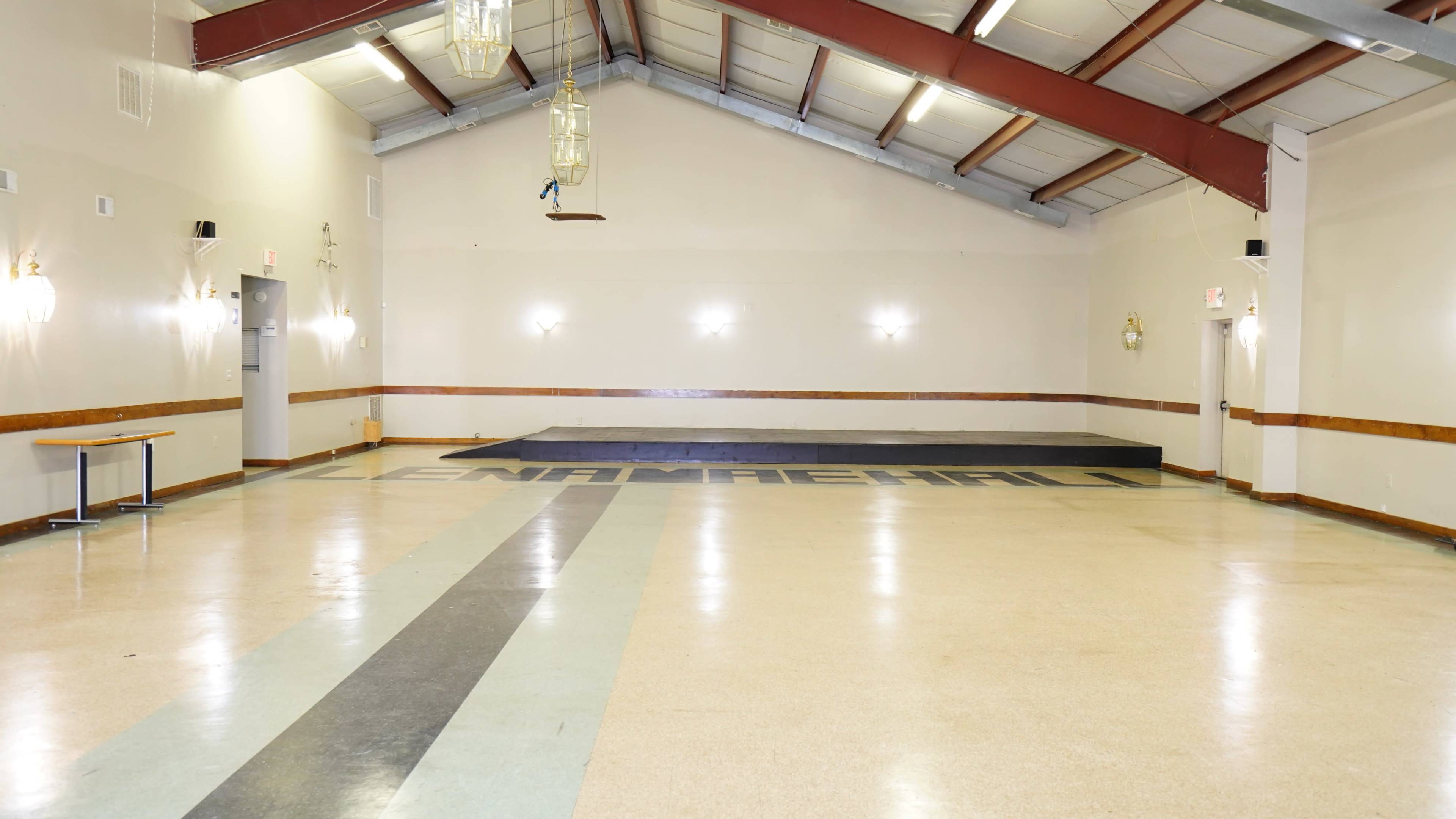 An empty hall with a wooden stage at one end and a large floor area featuring a dark stripe running down the center.
