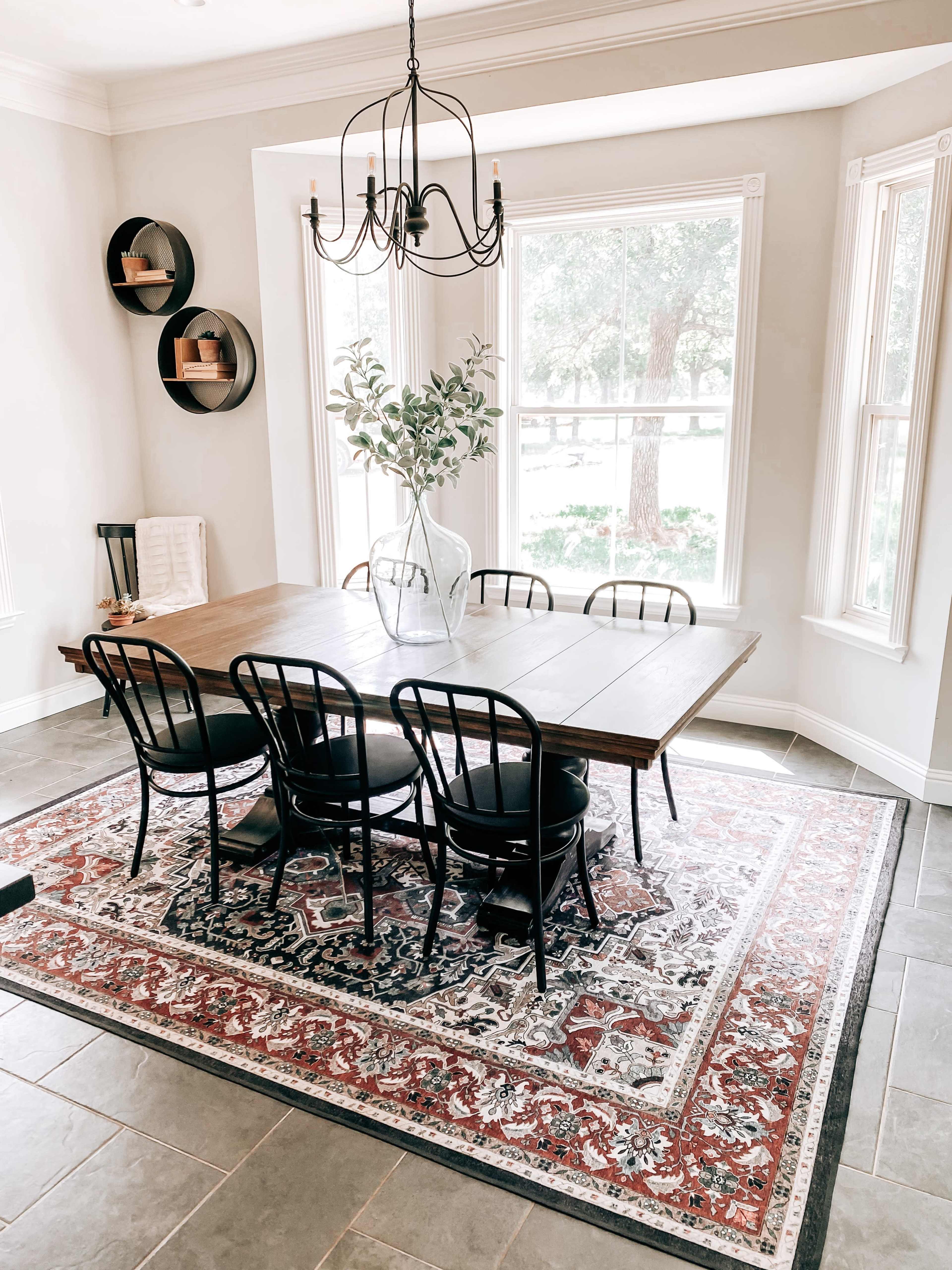A dining room features a wooden table surrounded by black chairs on a patterned rug, with large windows allowing natural light to fill the space.