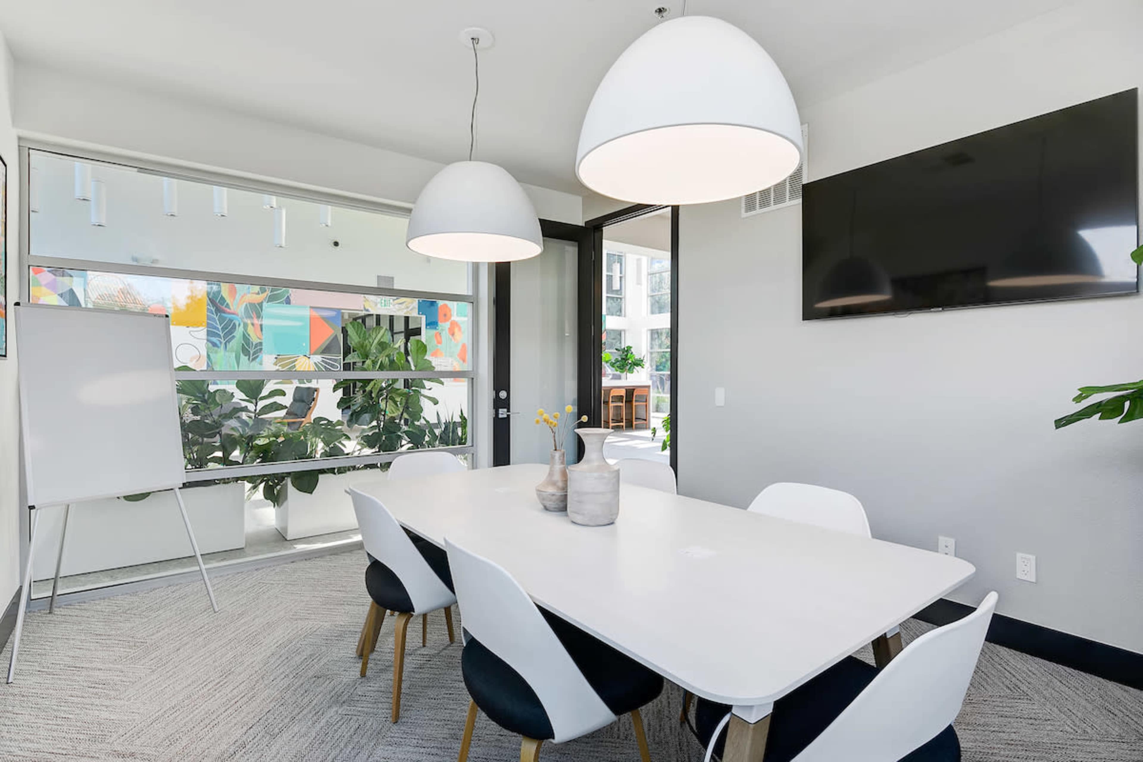 A modern conference room features a long white table surrounded by six chairs, with large pendant lights above and a television mounted on the wall.