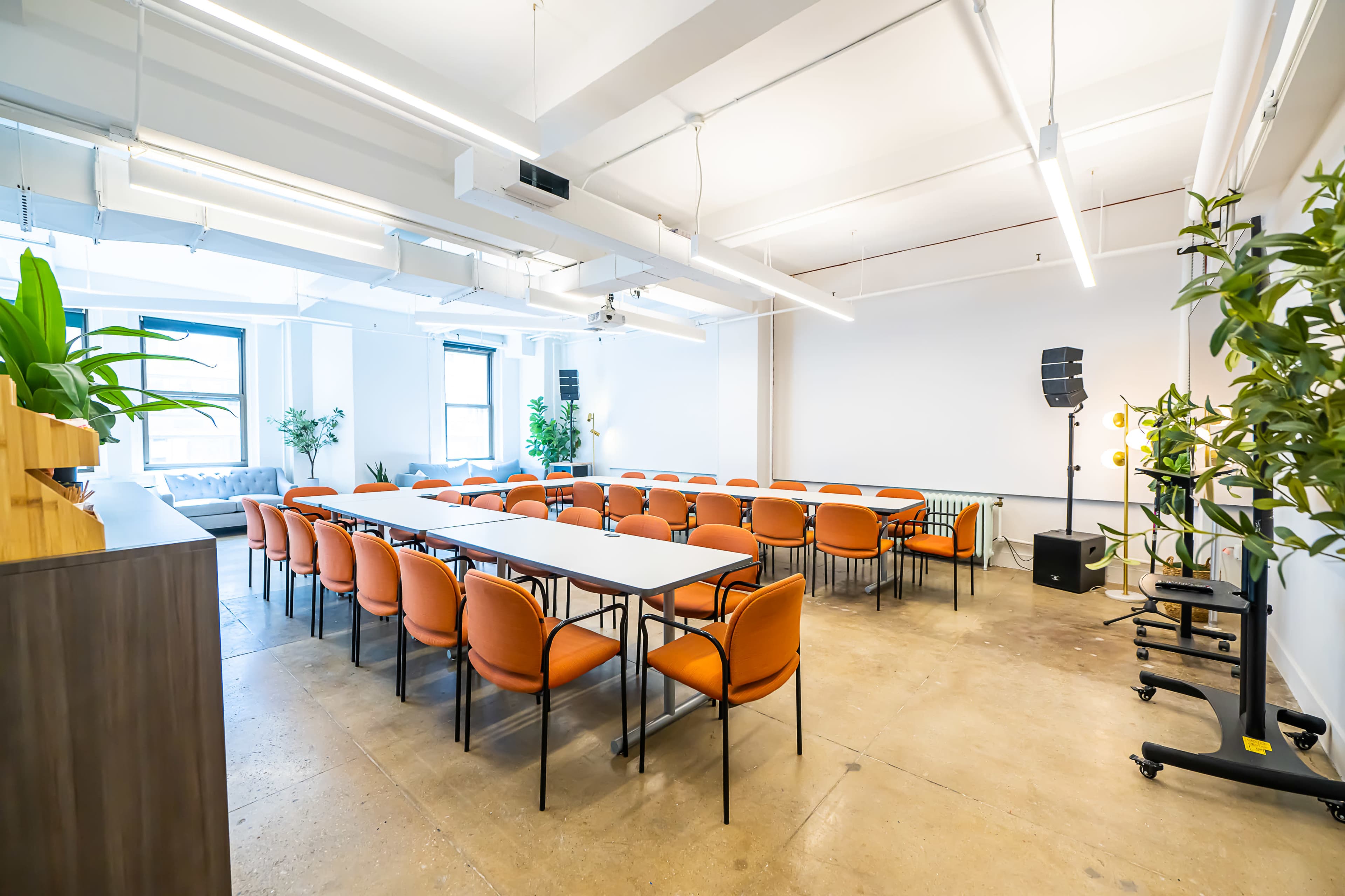 A modern conference room arranged with orange chairs around multiple tables, featuring large windows and potted plants.