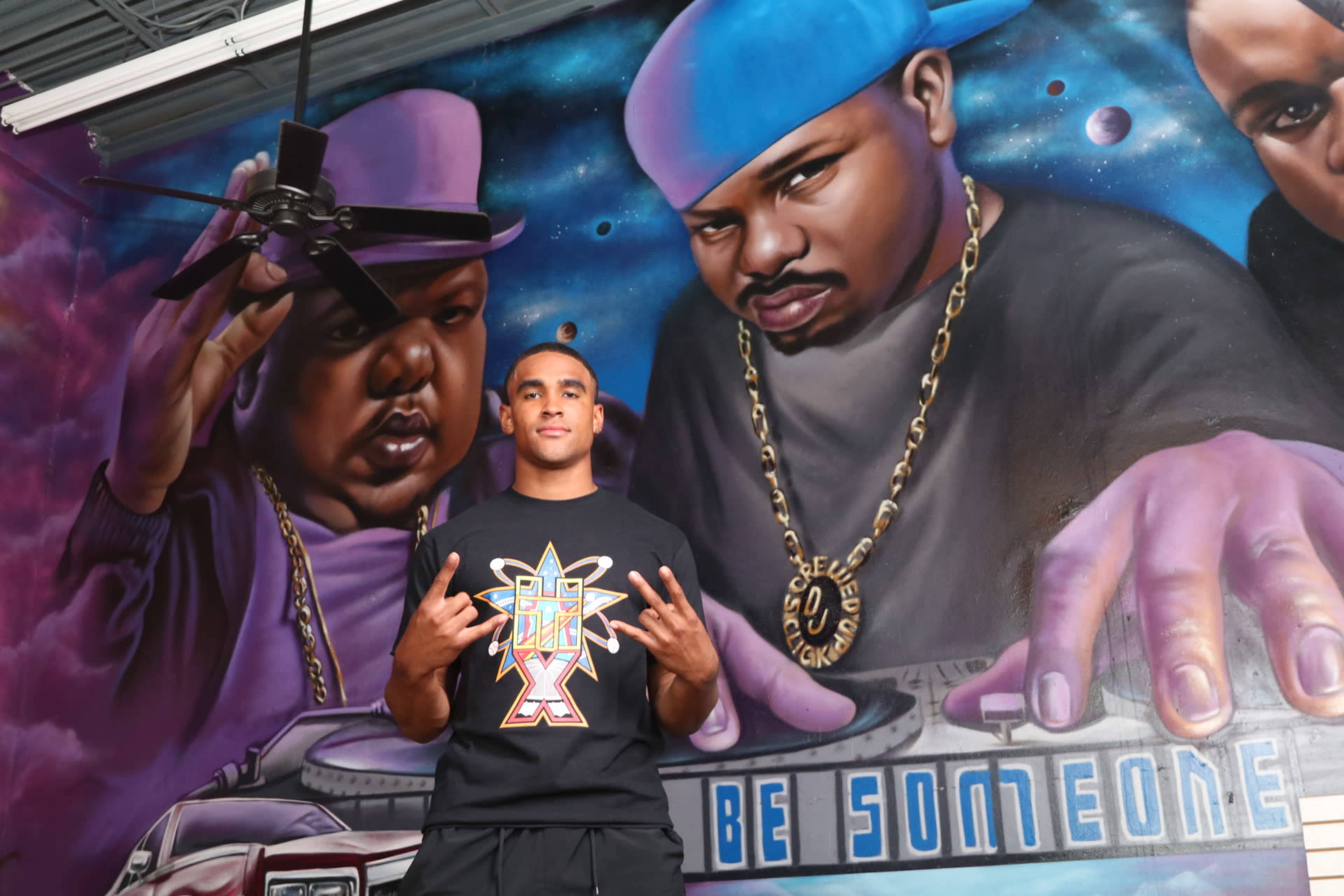 A young man poses in front of a colorful mural featuring two figures, one wearing a purple hat and the other a blue cap, beside a turntable with the words "BE SOMEONE" displayed.