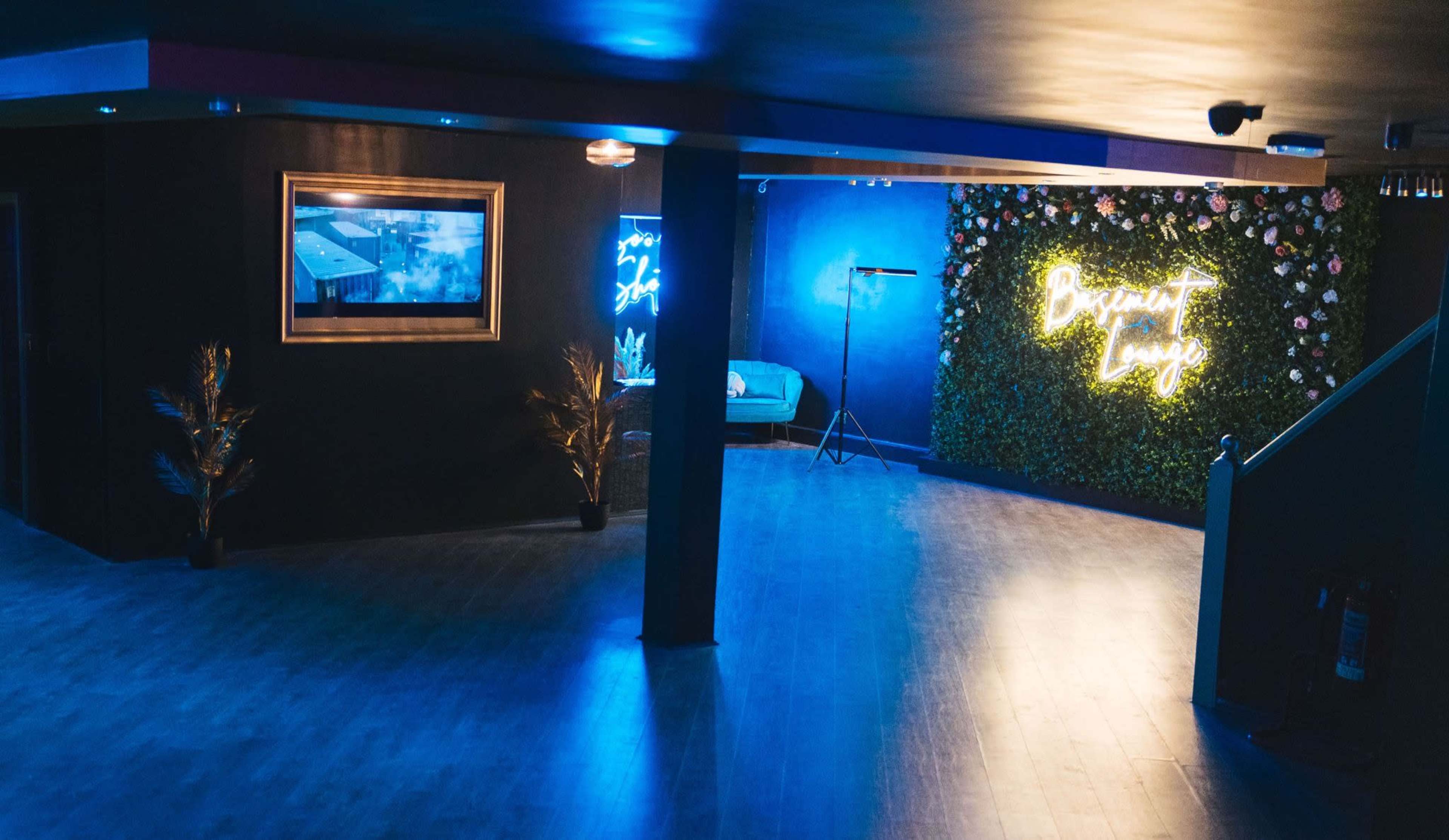The image shows a dimly lit interior space featuring a large green wall with neon signage, a framed picture on the wall, and a small seating area.