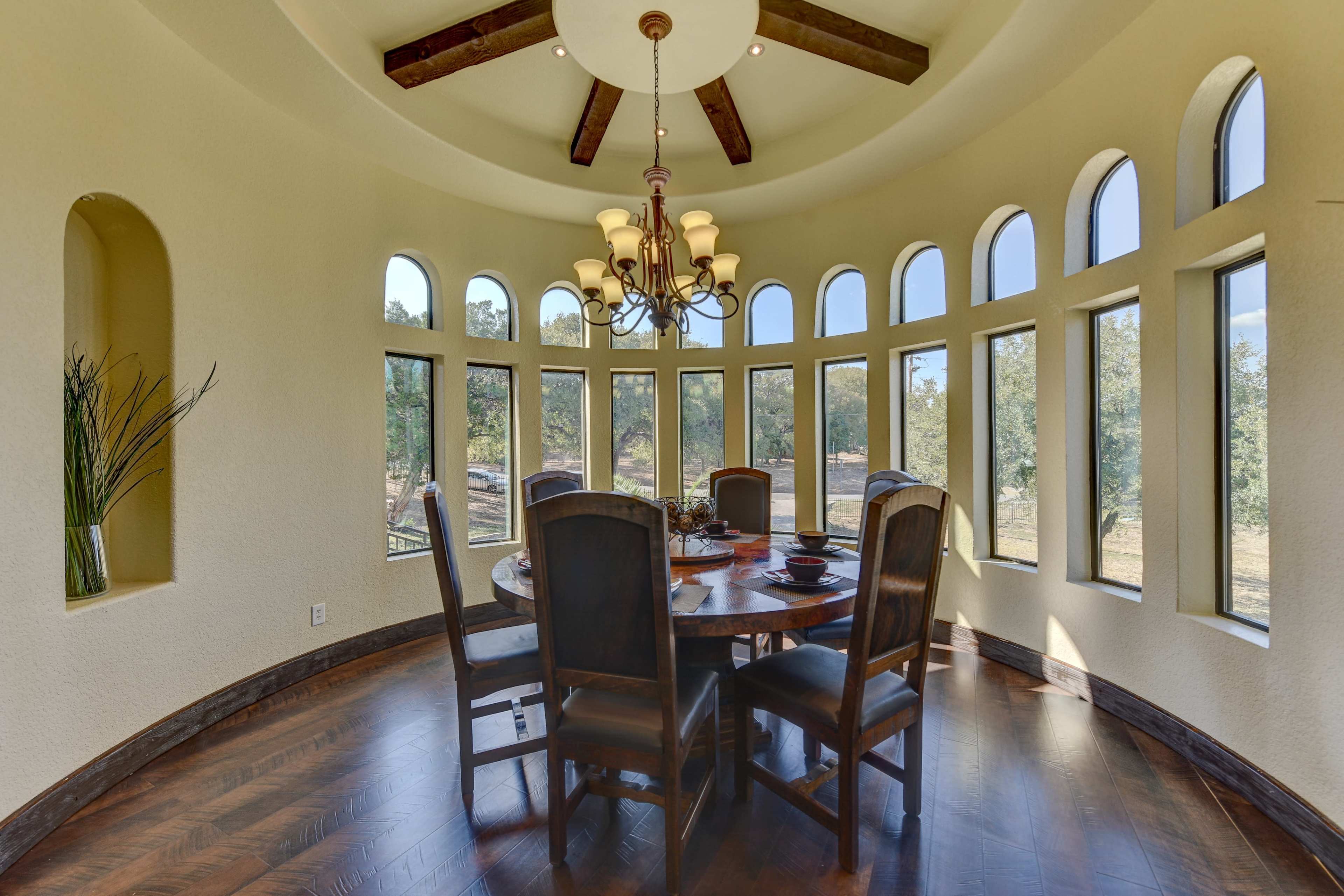 A circular dining room features a large wooden table surrounded by dark chairs, illuminated by a chandelier and natural light streaming through multiple arched windows.