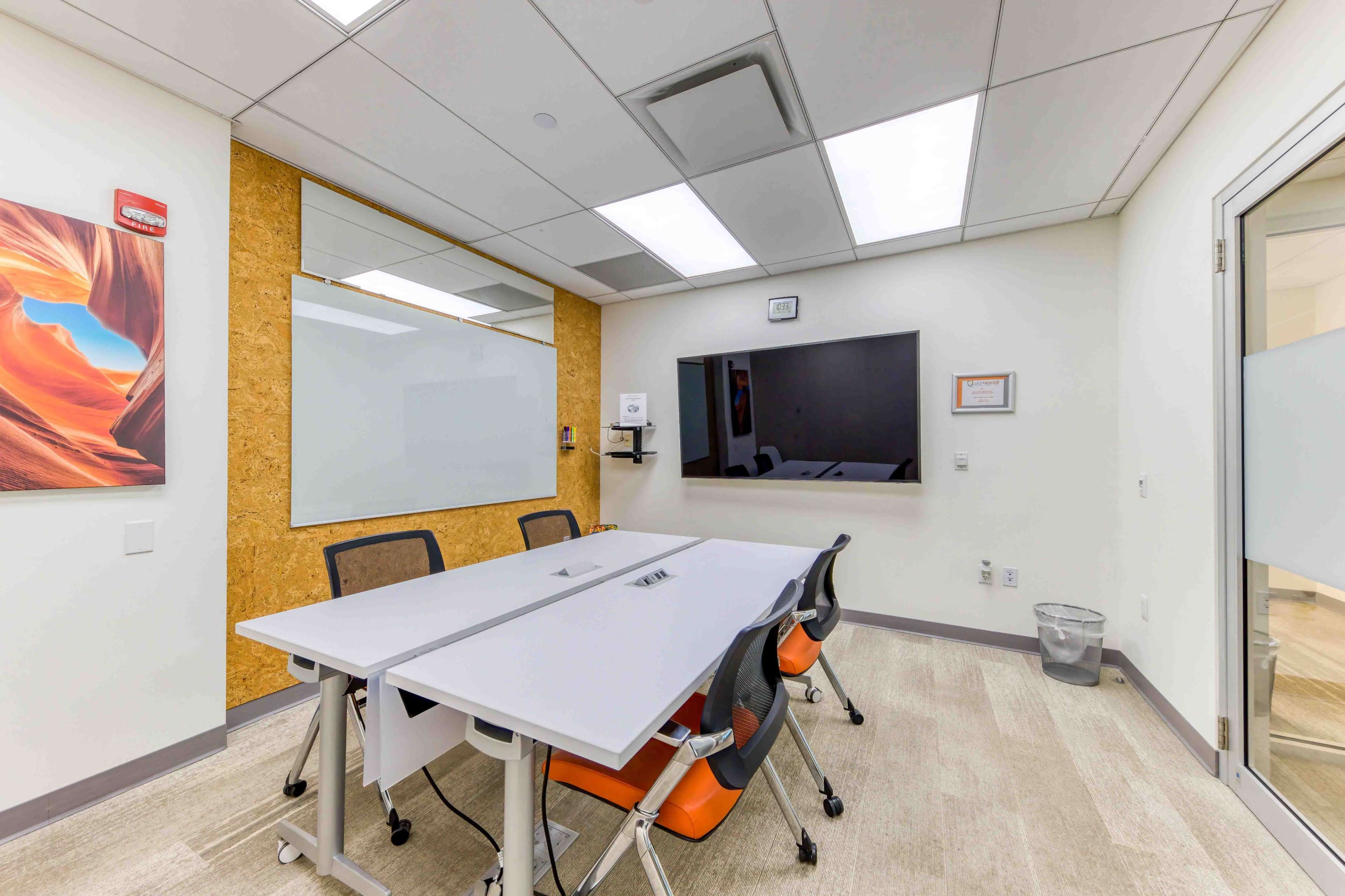 A conference room features a white table surrounded by orange and gray chairs, a wall-mounted screen, and a whiteboard.