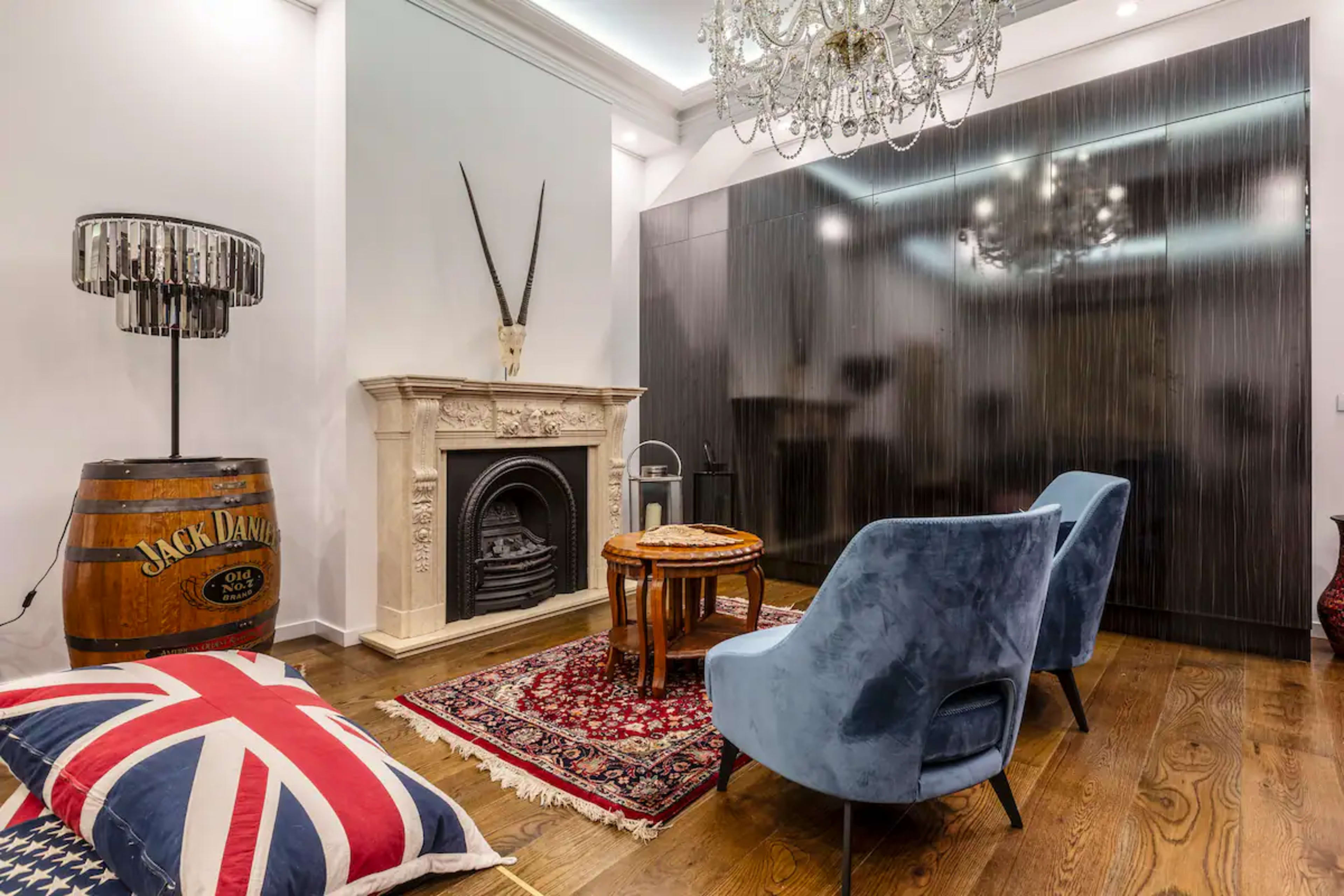 A modern living space featuring a decorative fireplace, blue armchairs, a wooden table, a patterned rug, and a vintage-style lamp beside a barrel.