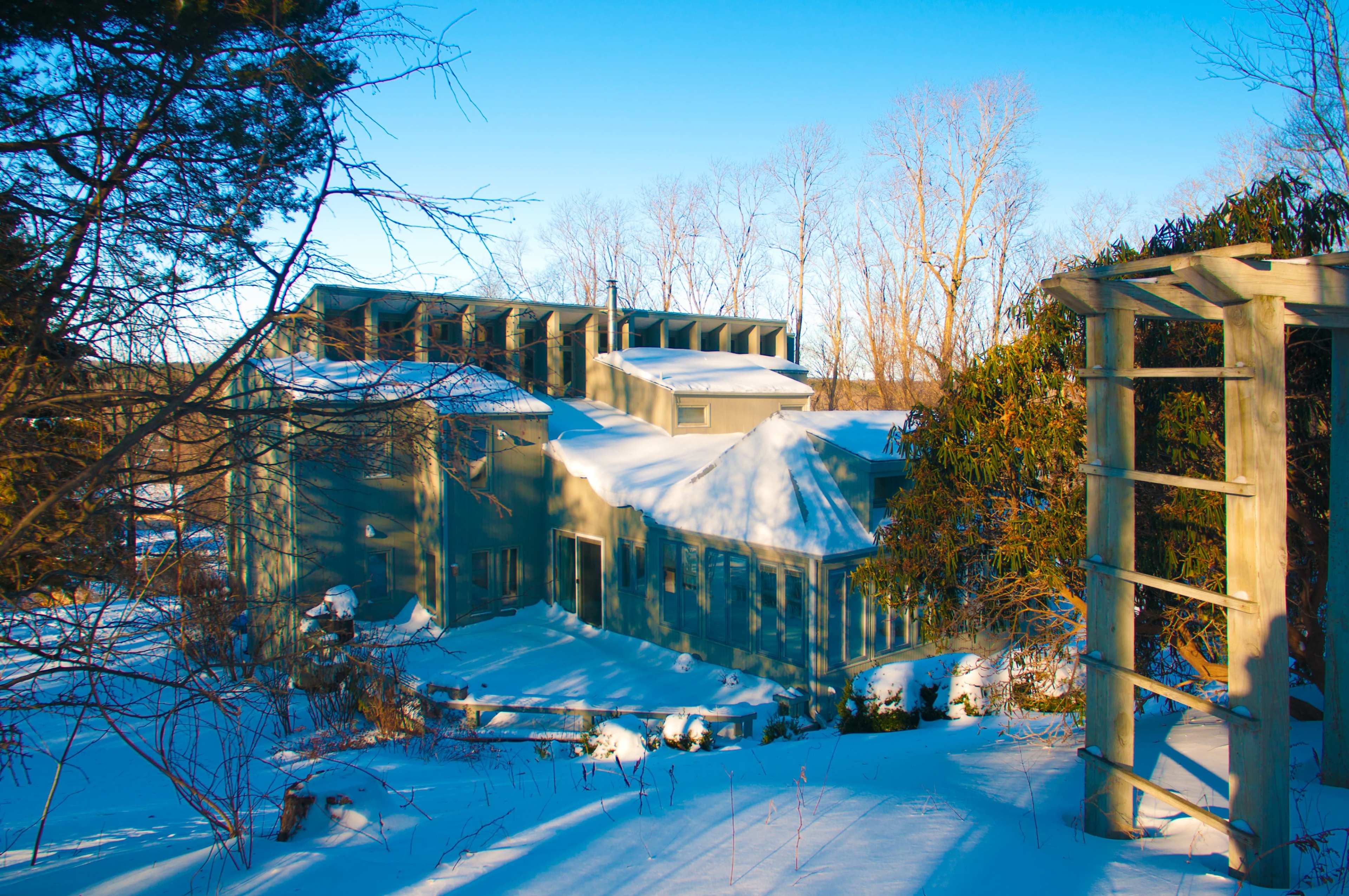 A modern, angular house is partially covered in snow and surrounded by leafless trees in a winter landscape.