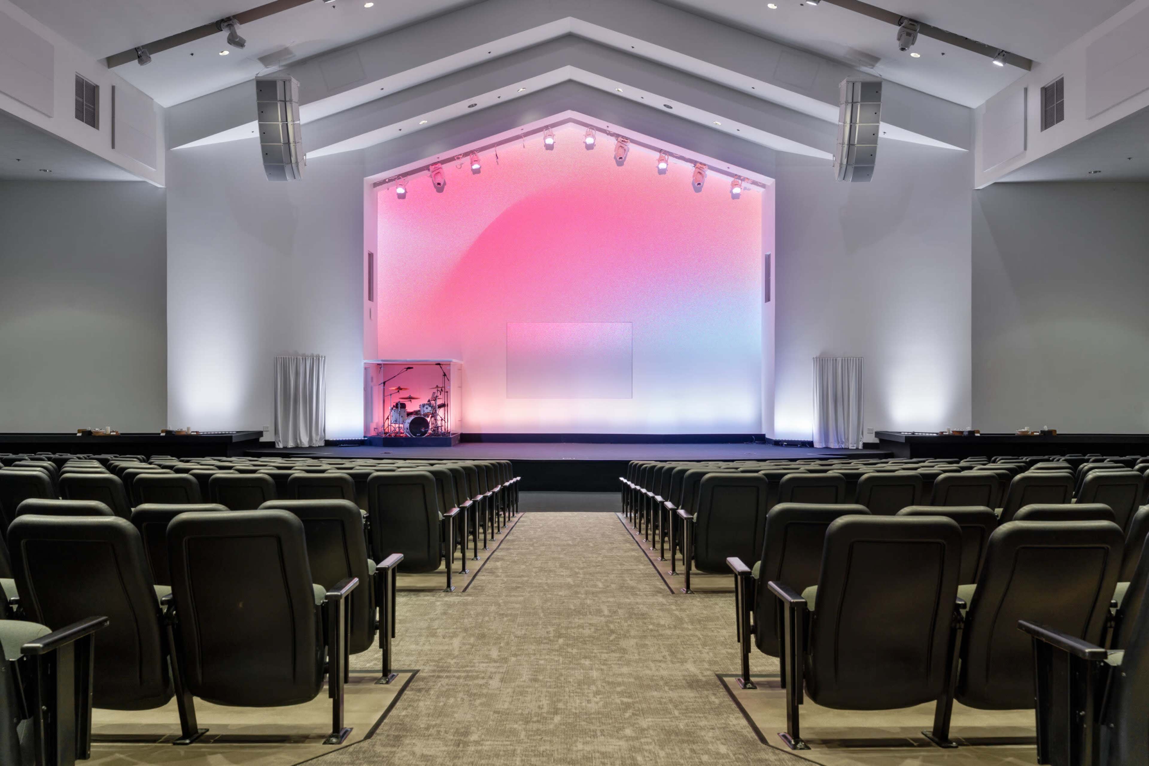 The image shows an empty auditorium with rows of black chairs facing a stage featuring colored lighting and a drum set in the background.