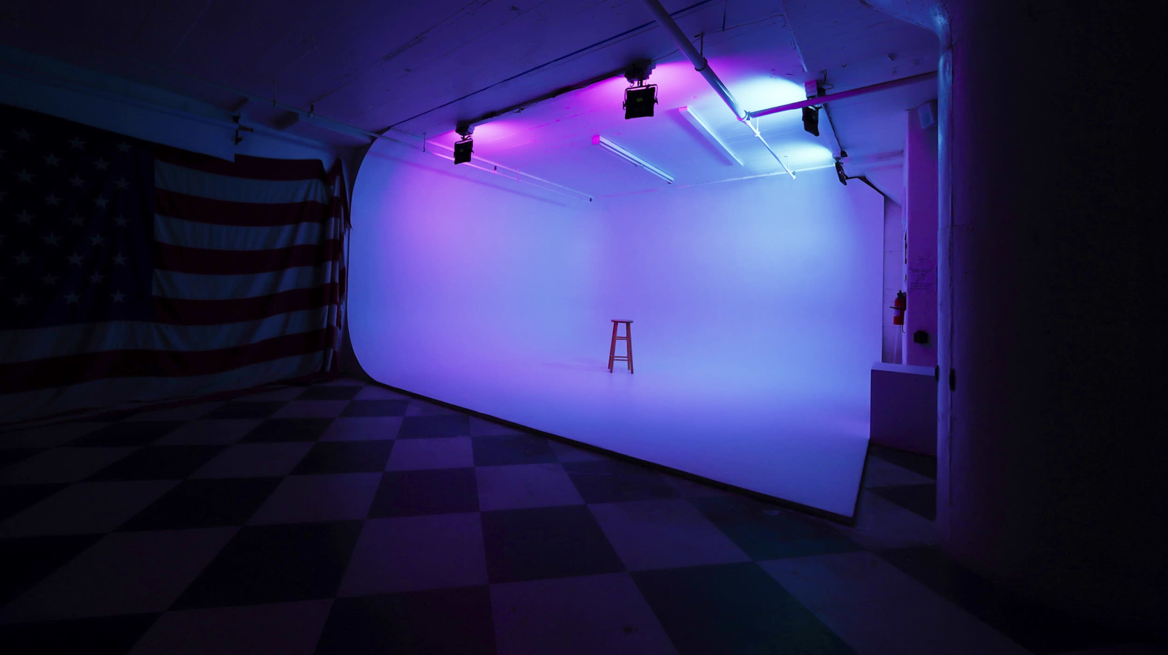 An empty photography studio features a single stool on a seamless backdrop, illuminated by colored lights, with an American flag draped on the wall.