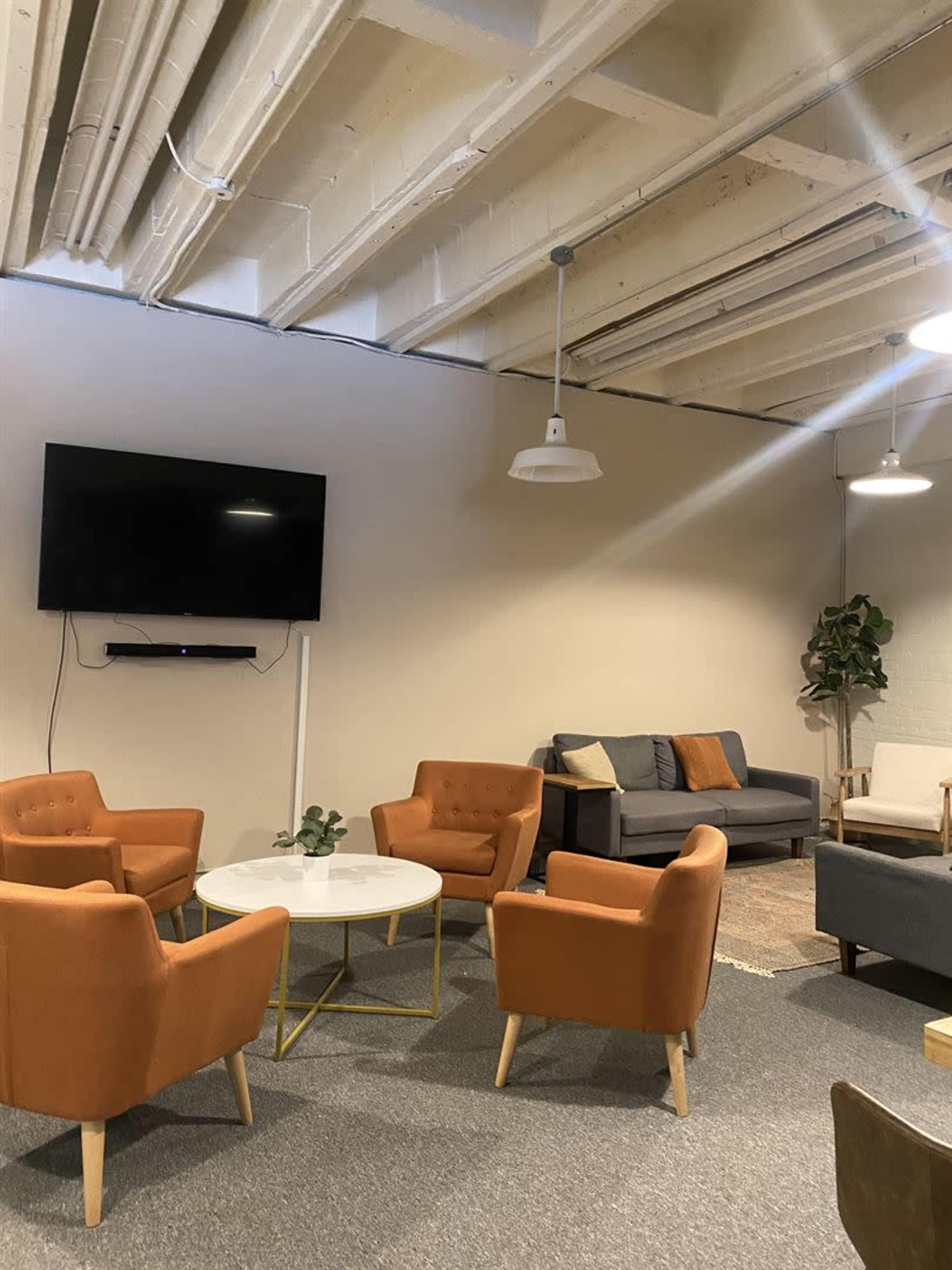 The image shows a lounge area with four orange chairs surrounding a circular coffee table, a gray couch against the wall, and a mounted television above the seating.