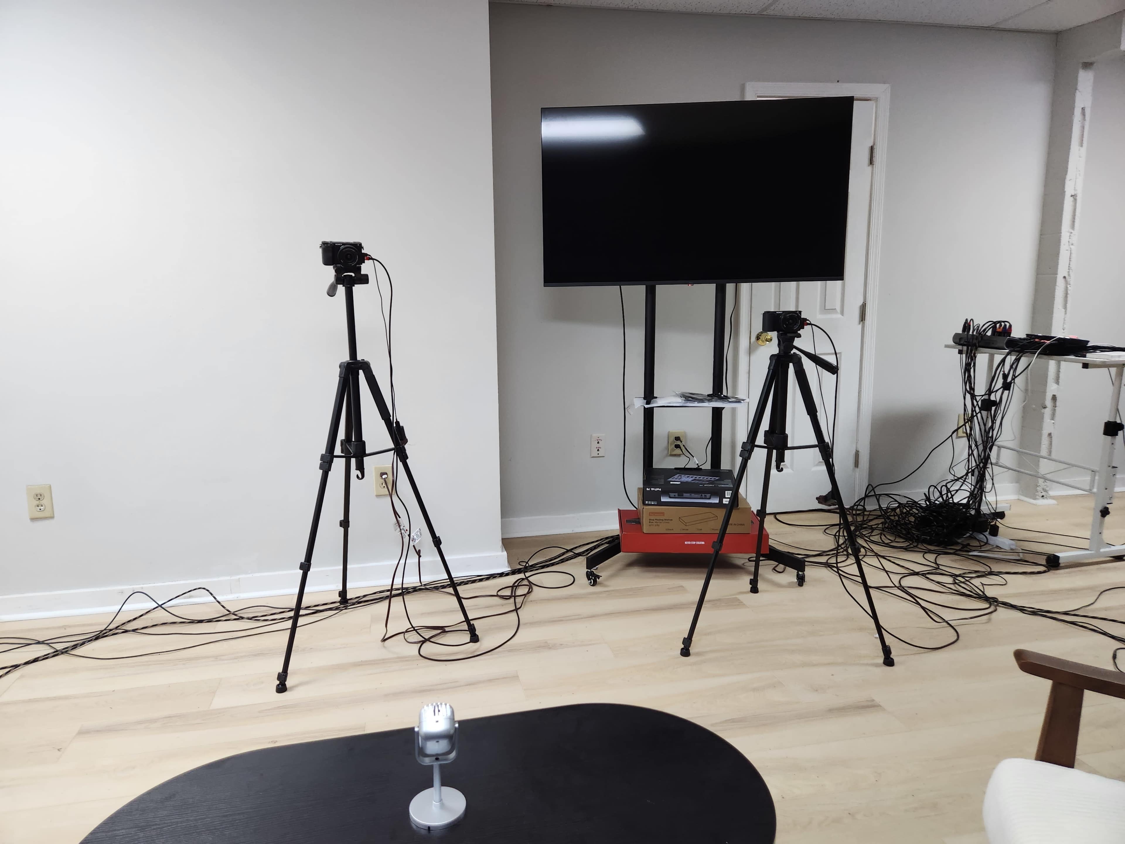 The image shows a room with two tripods and cameras positioned facing a television mounted on the wall, along with various tangled wires on the floor.