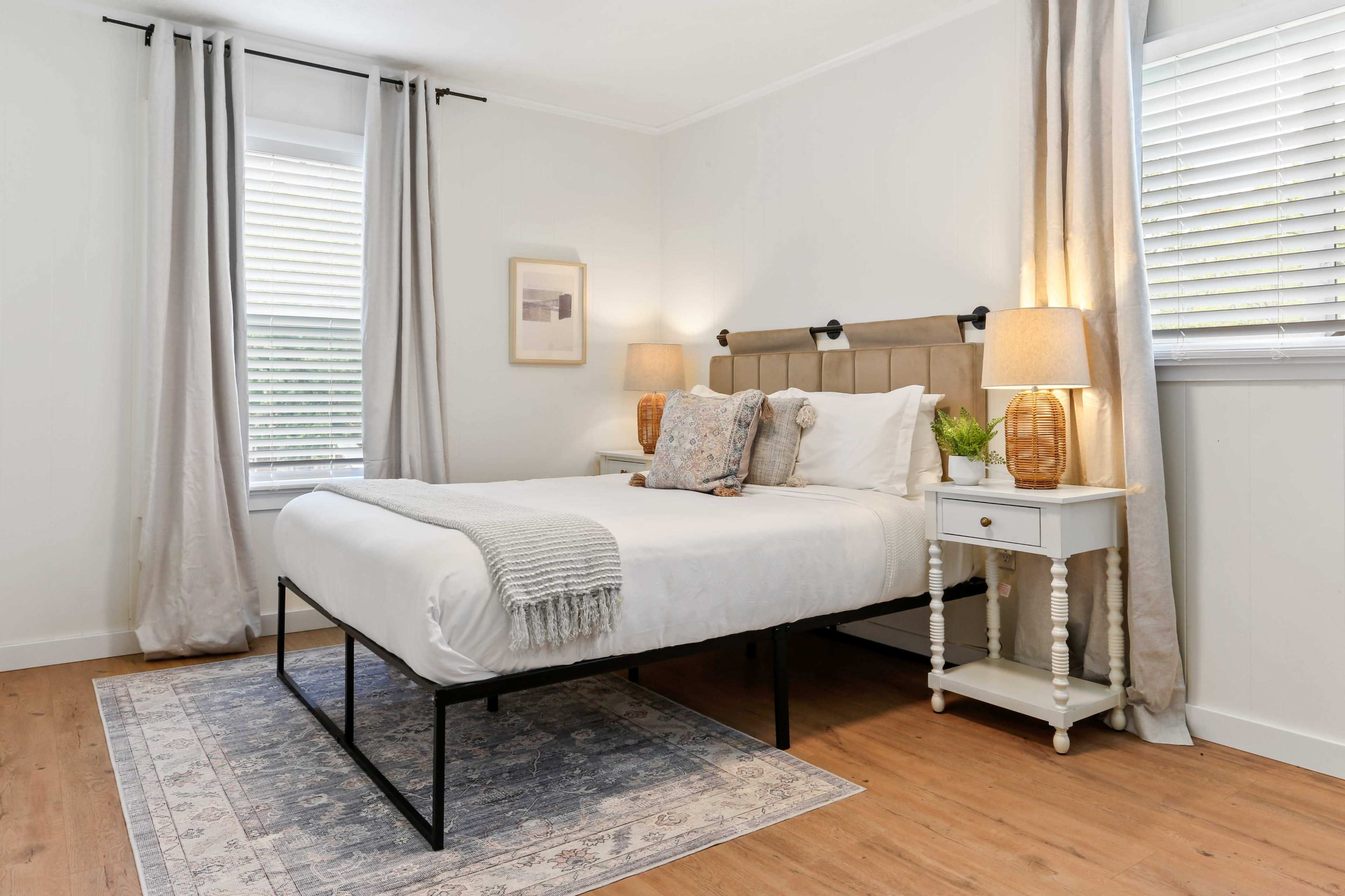 The image shows a neatly arranged bedroom with a bed, bedside tables, lamps, and large windows covered by curtains.