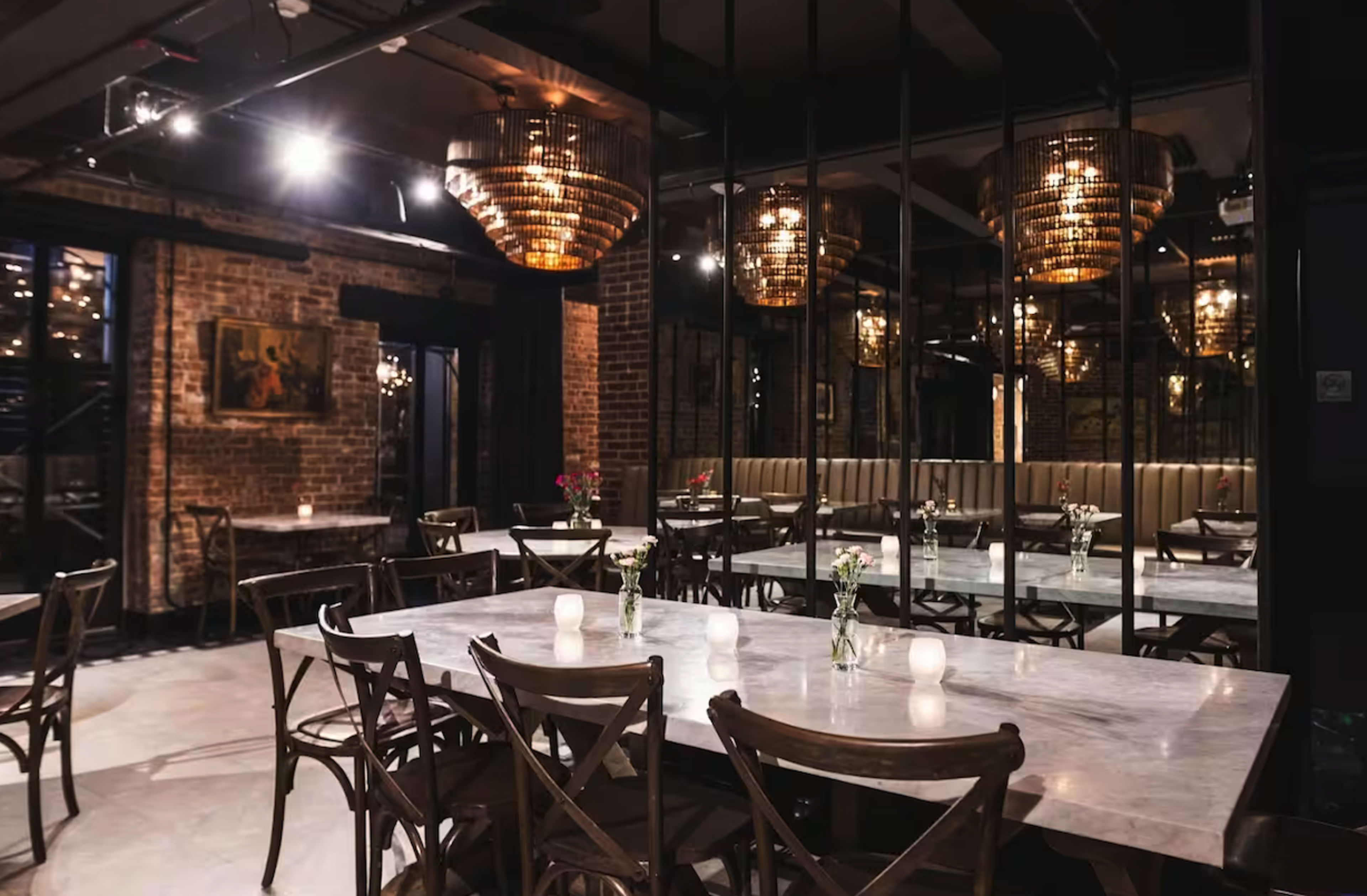 A modern restaurant interior features wooden tables, chairs, and large, ornate chandeliers, with exposed brick walls and mirrors reflecting the space.
