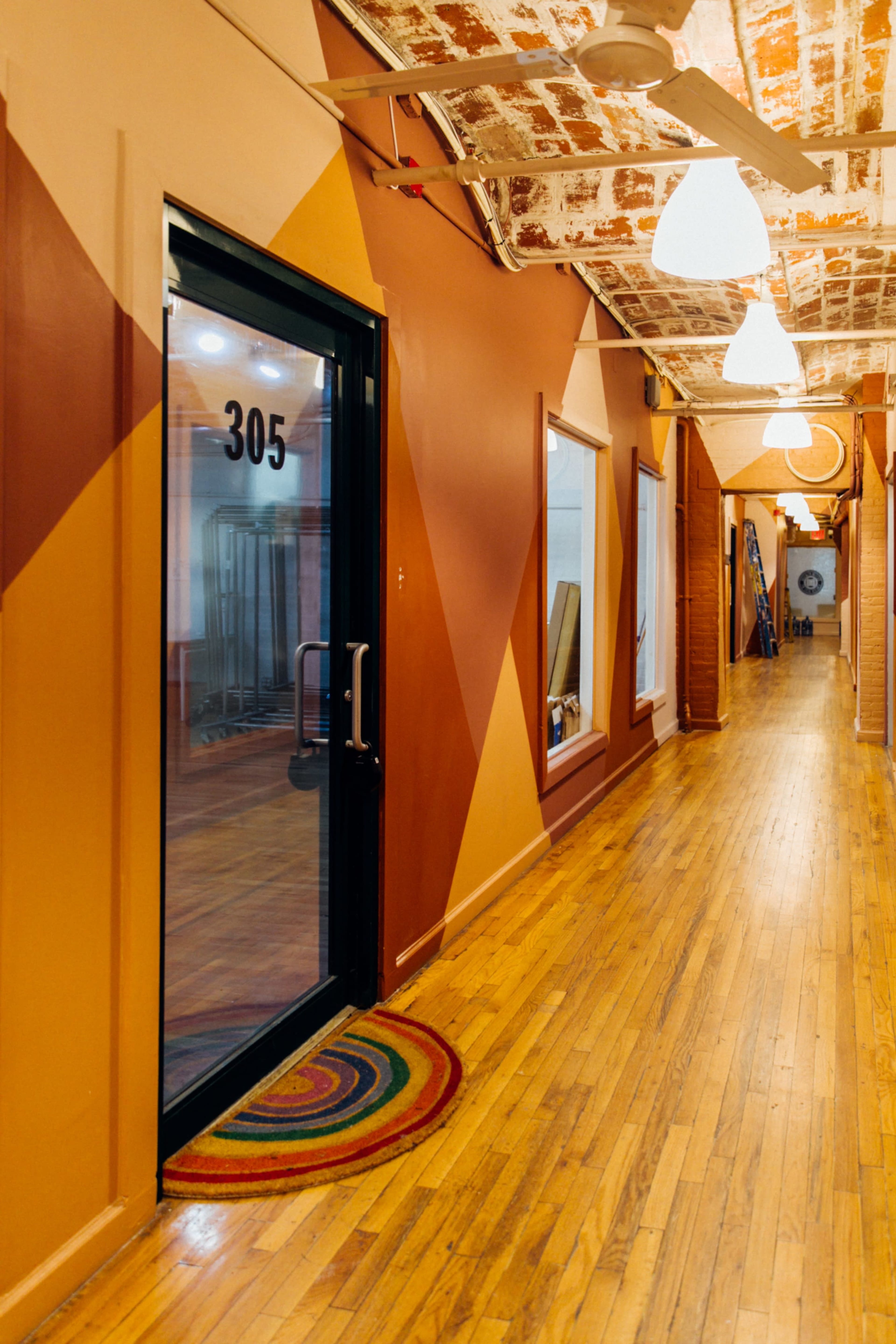 A narrow hallway featuring wooden floors and brightly colored walls leads to a door marked "305," accompanied by a circular doormat at its entrance.