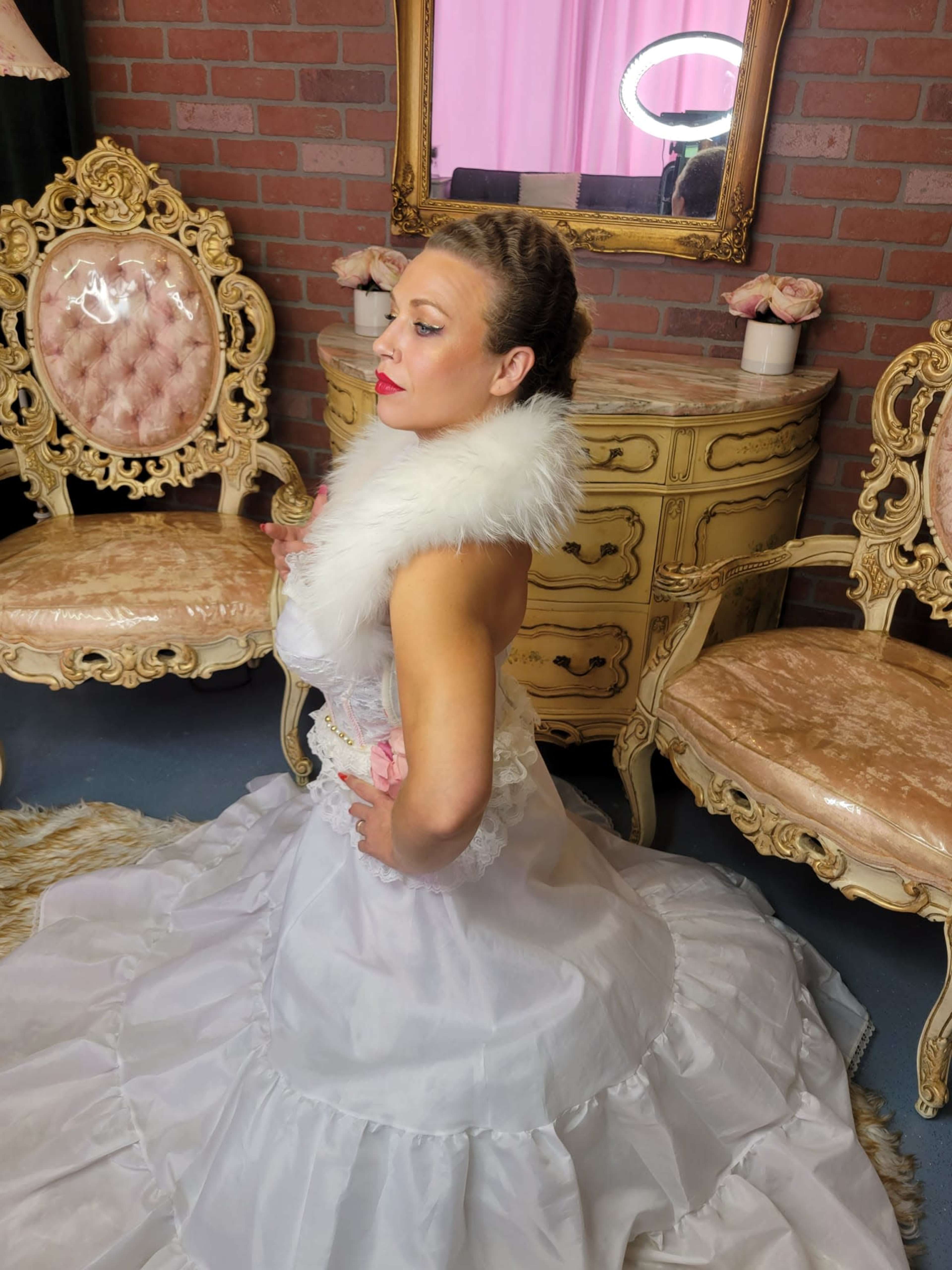 A woman in a white gown poses gracefully in a vintage-styled room with ornate furniture and a mirror in the background.