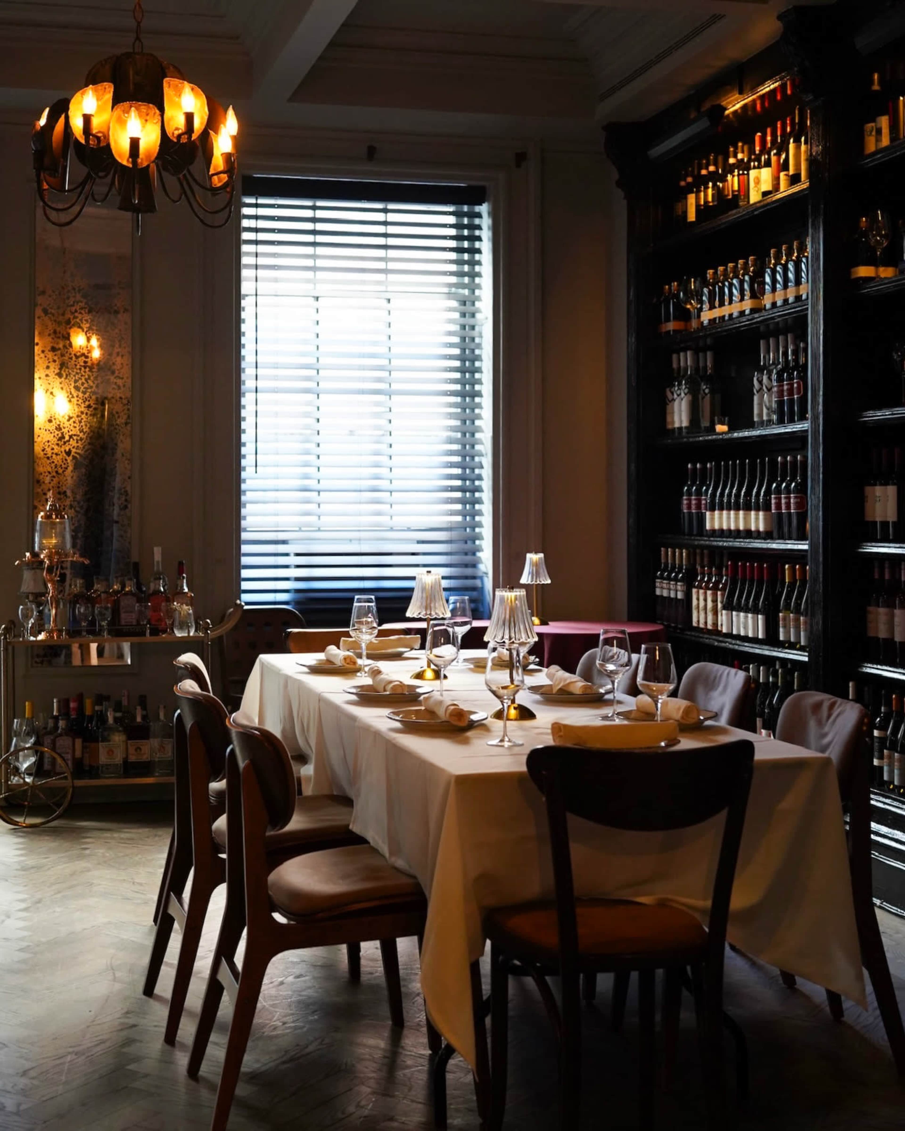 A elegantly set dining table with a white tablecloth is positioned near a window, surrounded by wooden chairs and a wall of wine bottles.