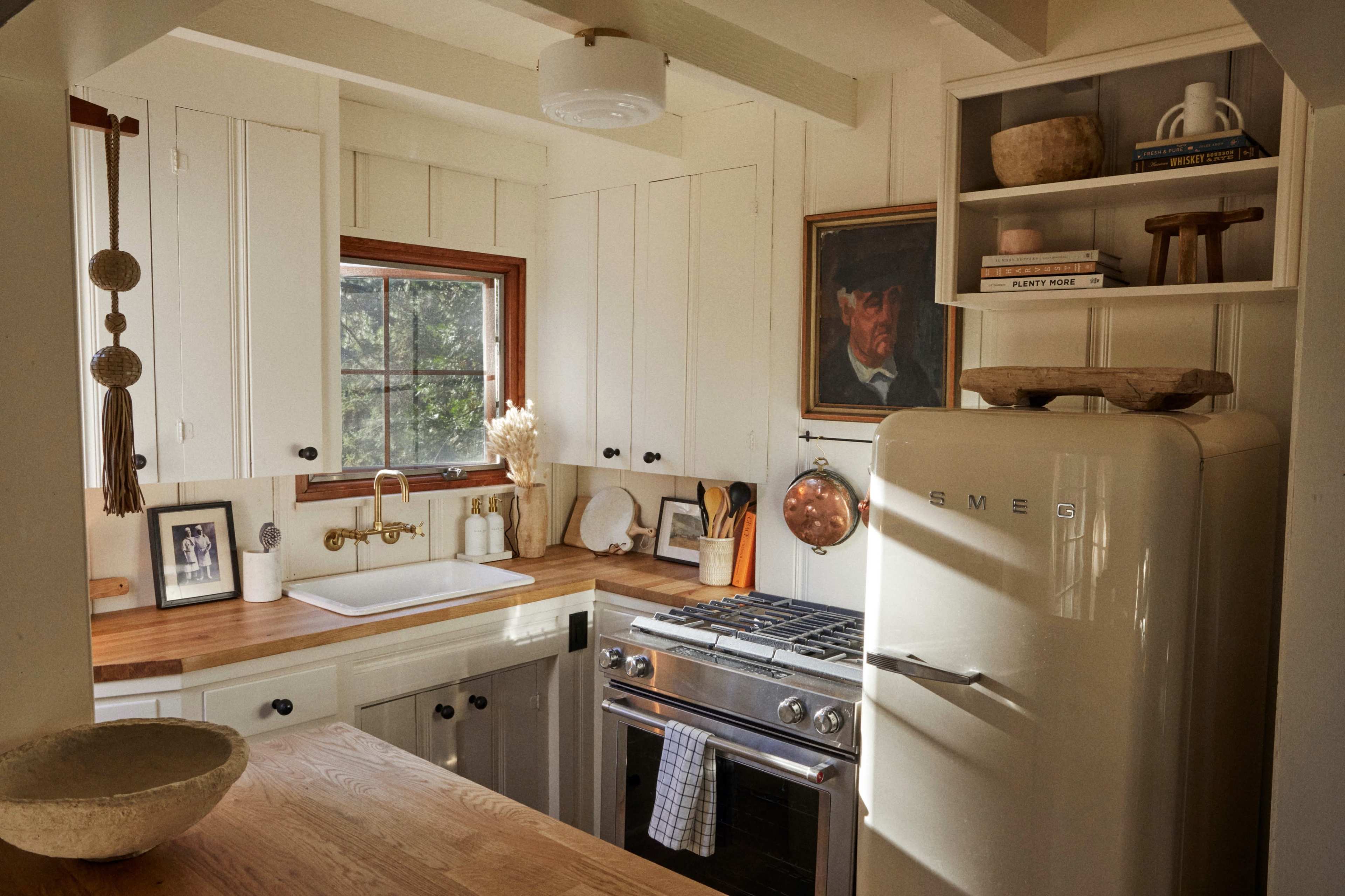 A cozy kitchen features white wooden cabinets, a stainless steel stove, a vintage fridge, and a window overlooking greenery.