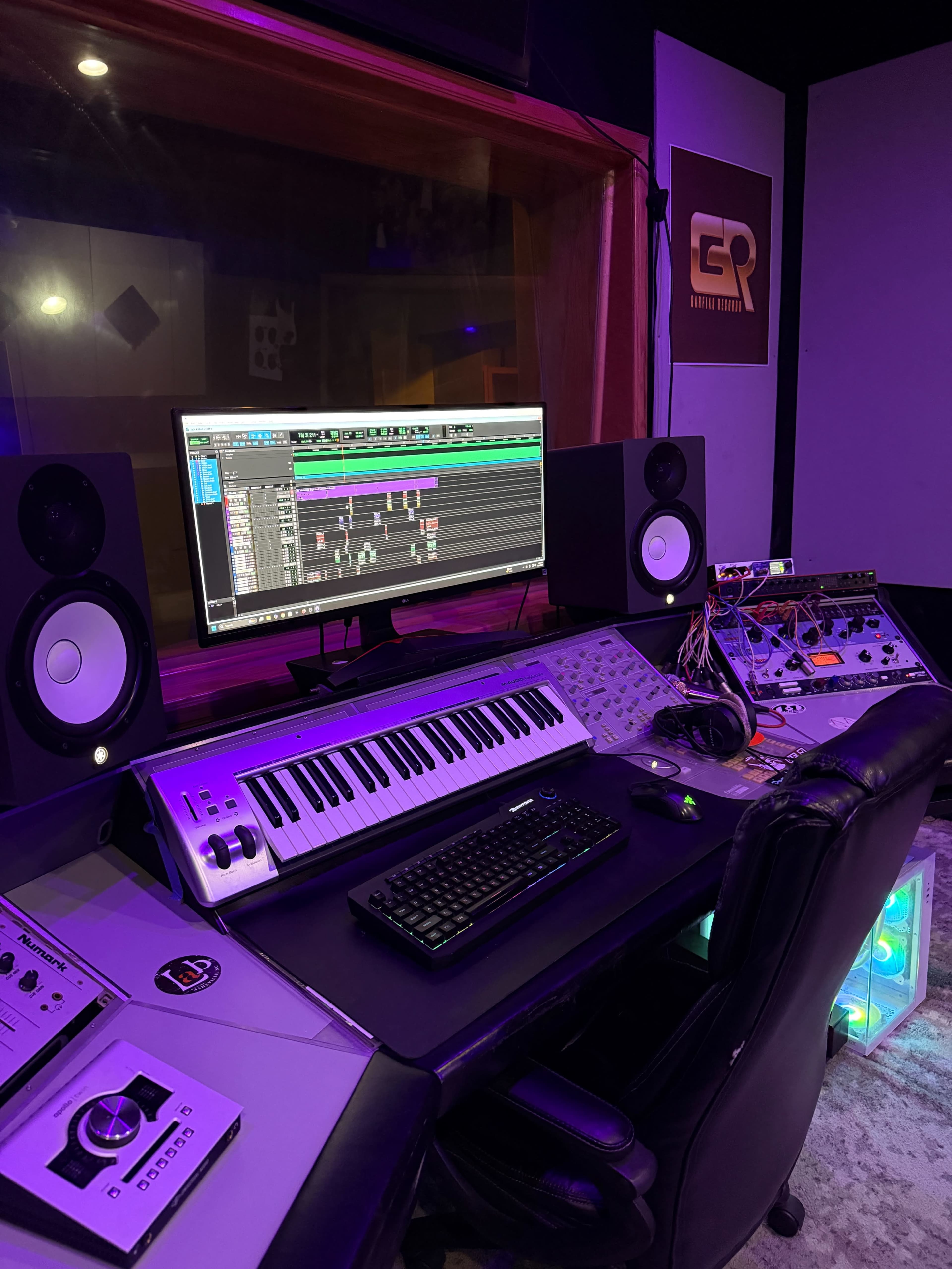 The image shows a music production studio featuring a keyboard, audio interface, computer monitor displaying music software, and studio monitors.