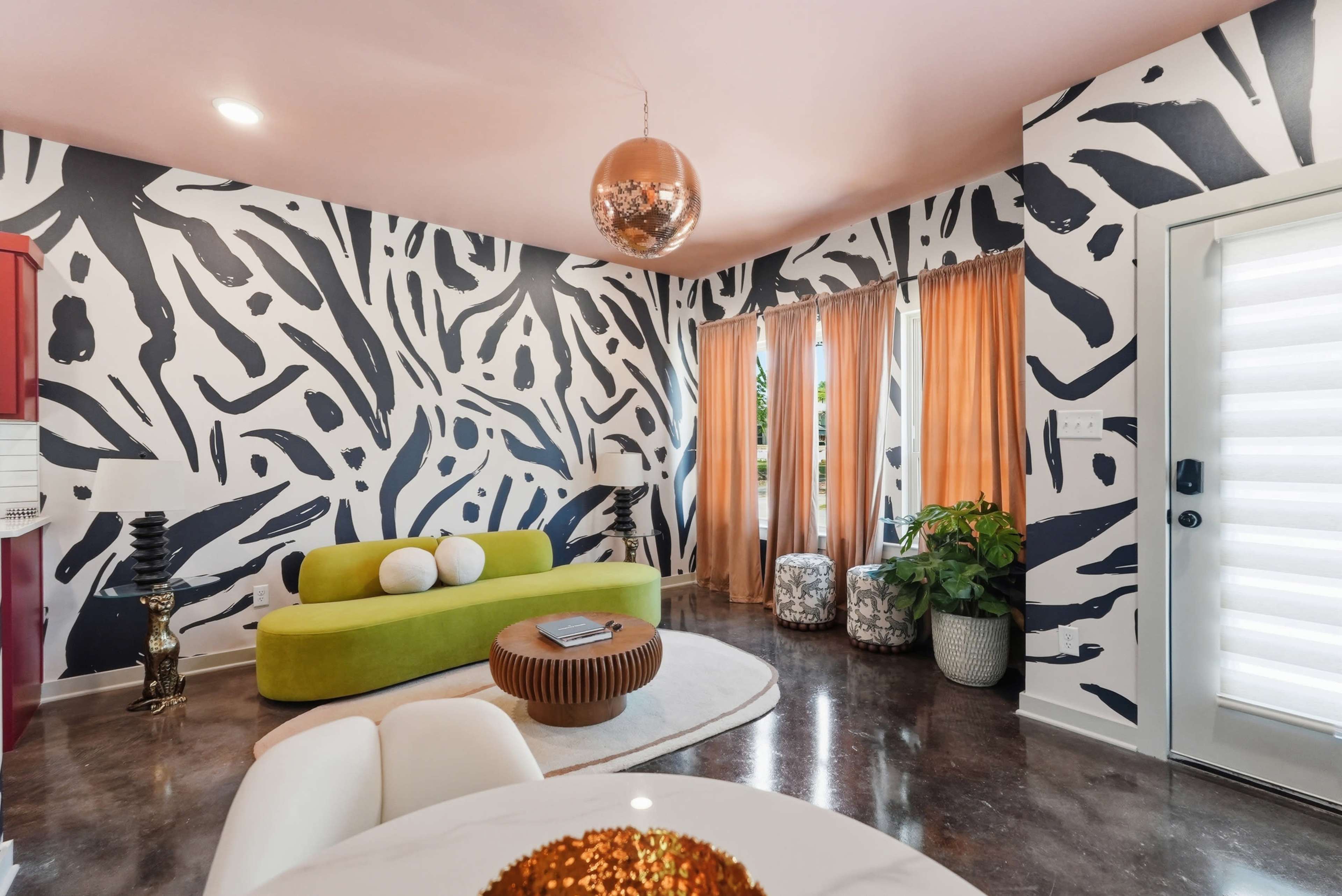 The room features an abstract black-and-white patterned wall, a vibrant green sofa, and large windows covered with orange curtains.