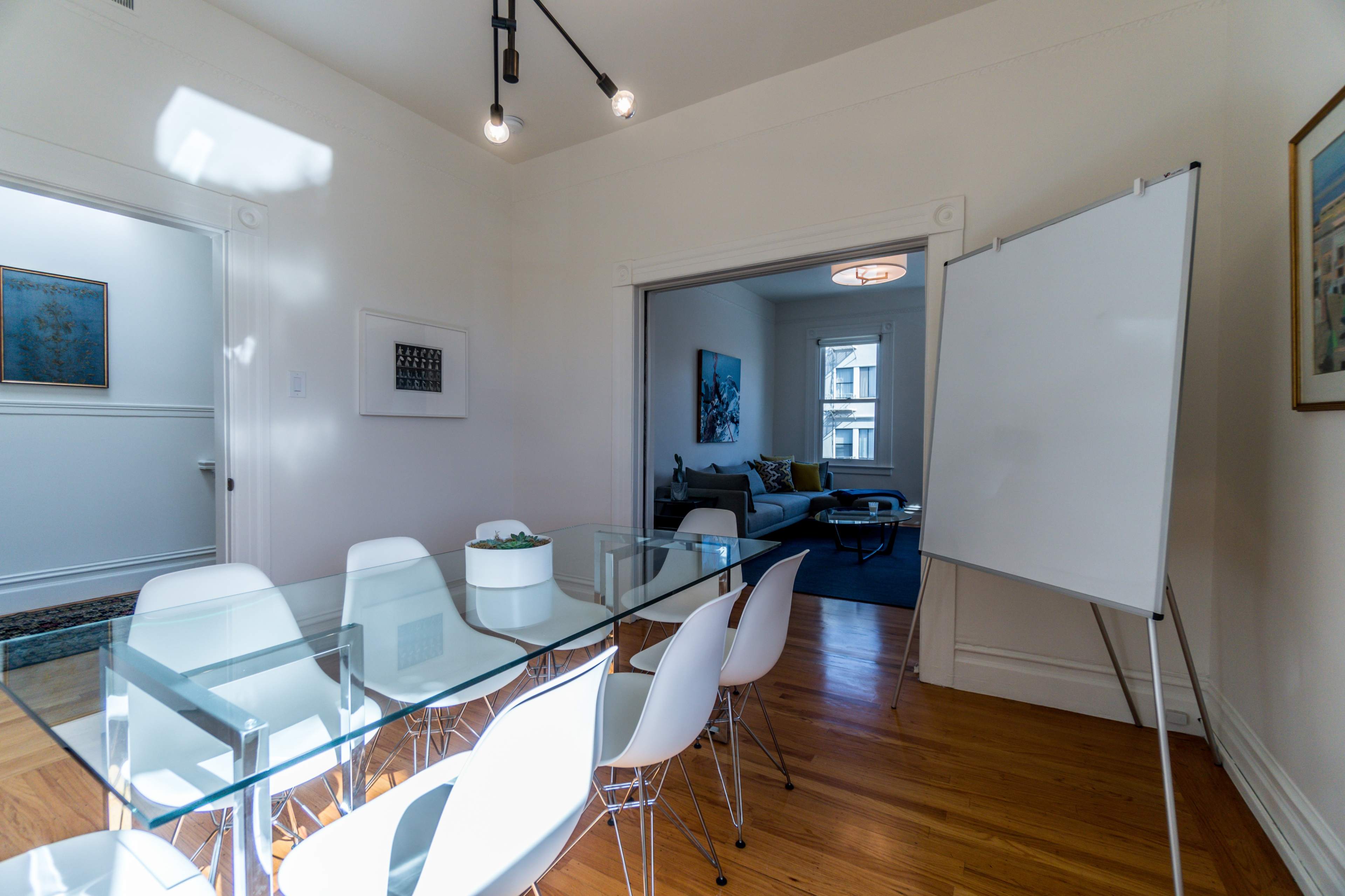 A modern conference room with a glass table, white chairs, and a flip chart stands adjacent to a living area with a couch and artwork.