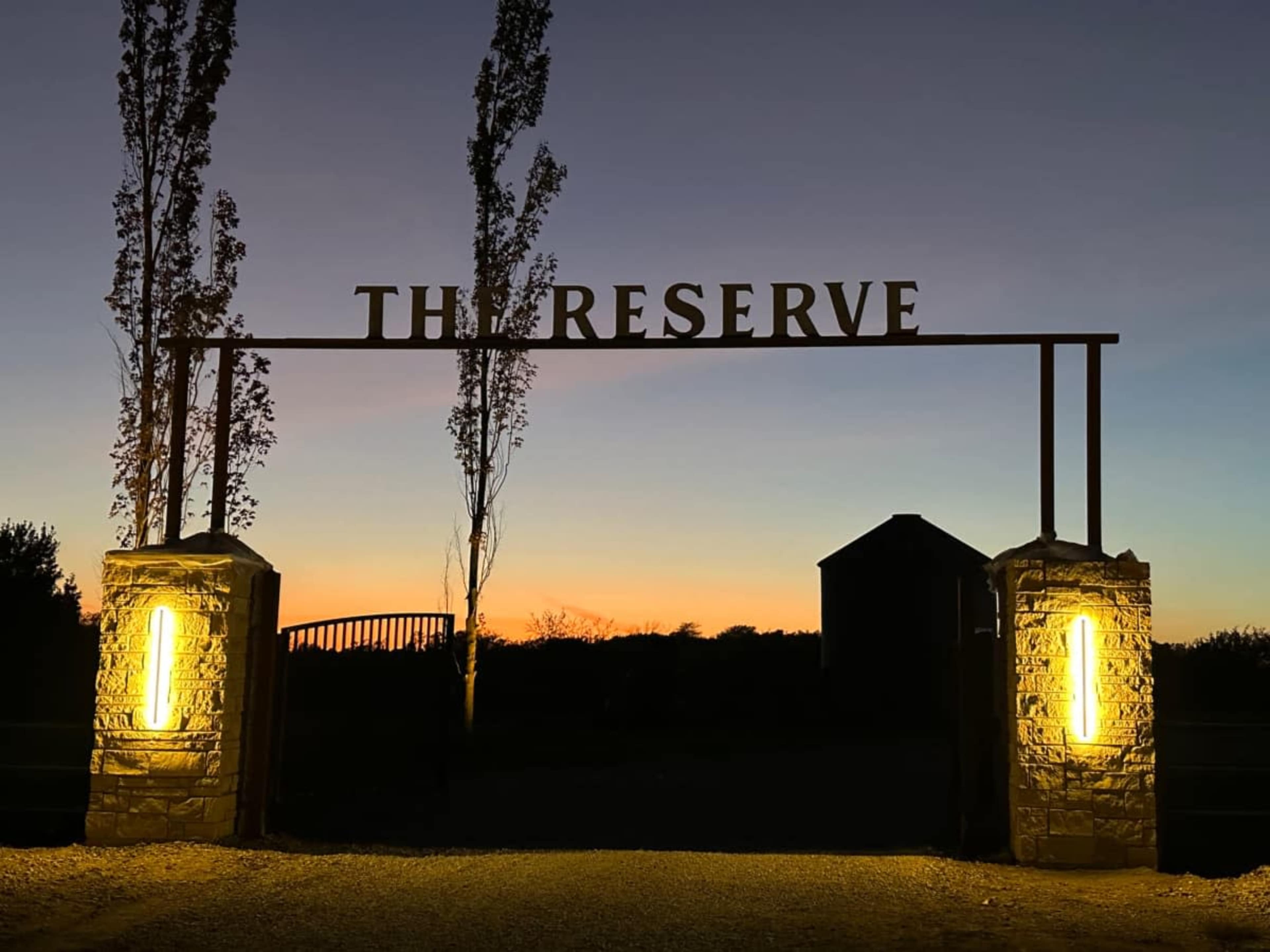 The image shows an entrance gate with the words "THE RESERVE" illuminated against a twilight sky, flanked by lit stone pillars and trees.