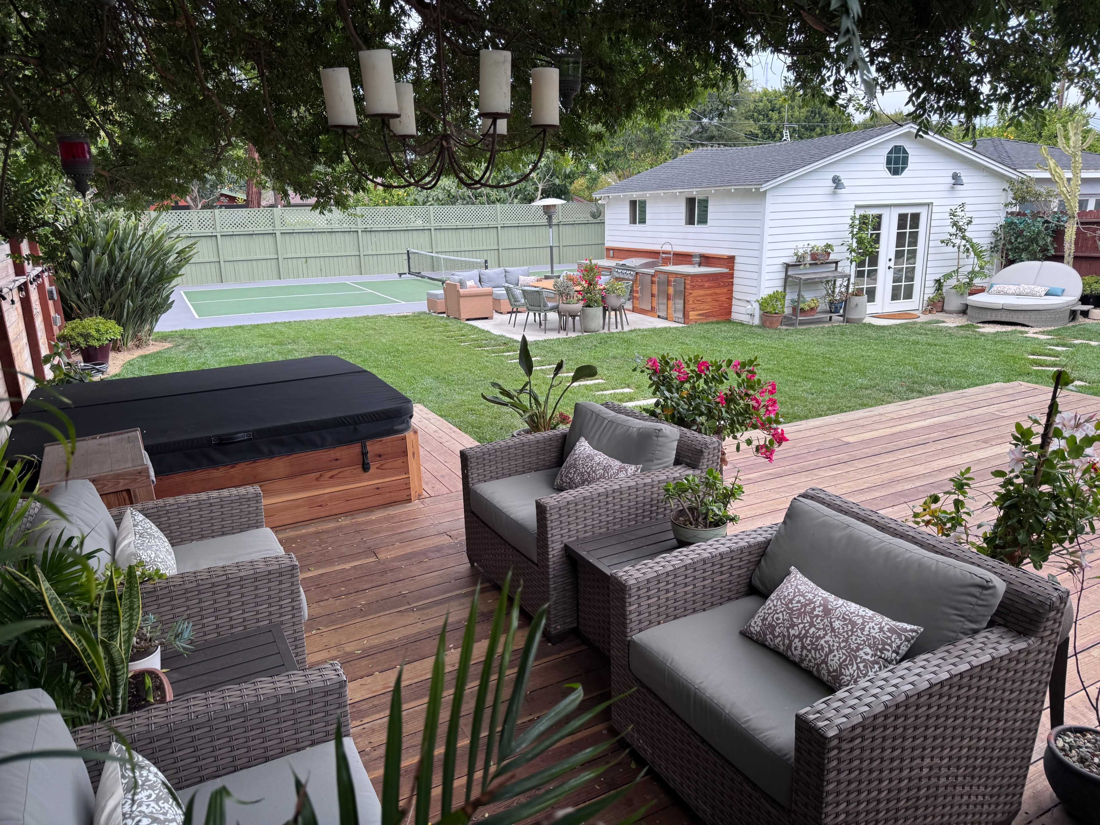 A backyard scene features a seating area with gray wicker furniture, a hot tub, and a well-maintained lawn with a tennis court and a garden shed in the background.
