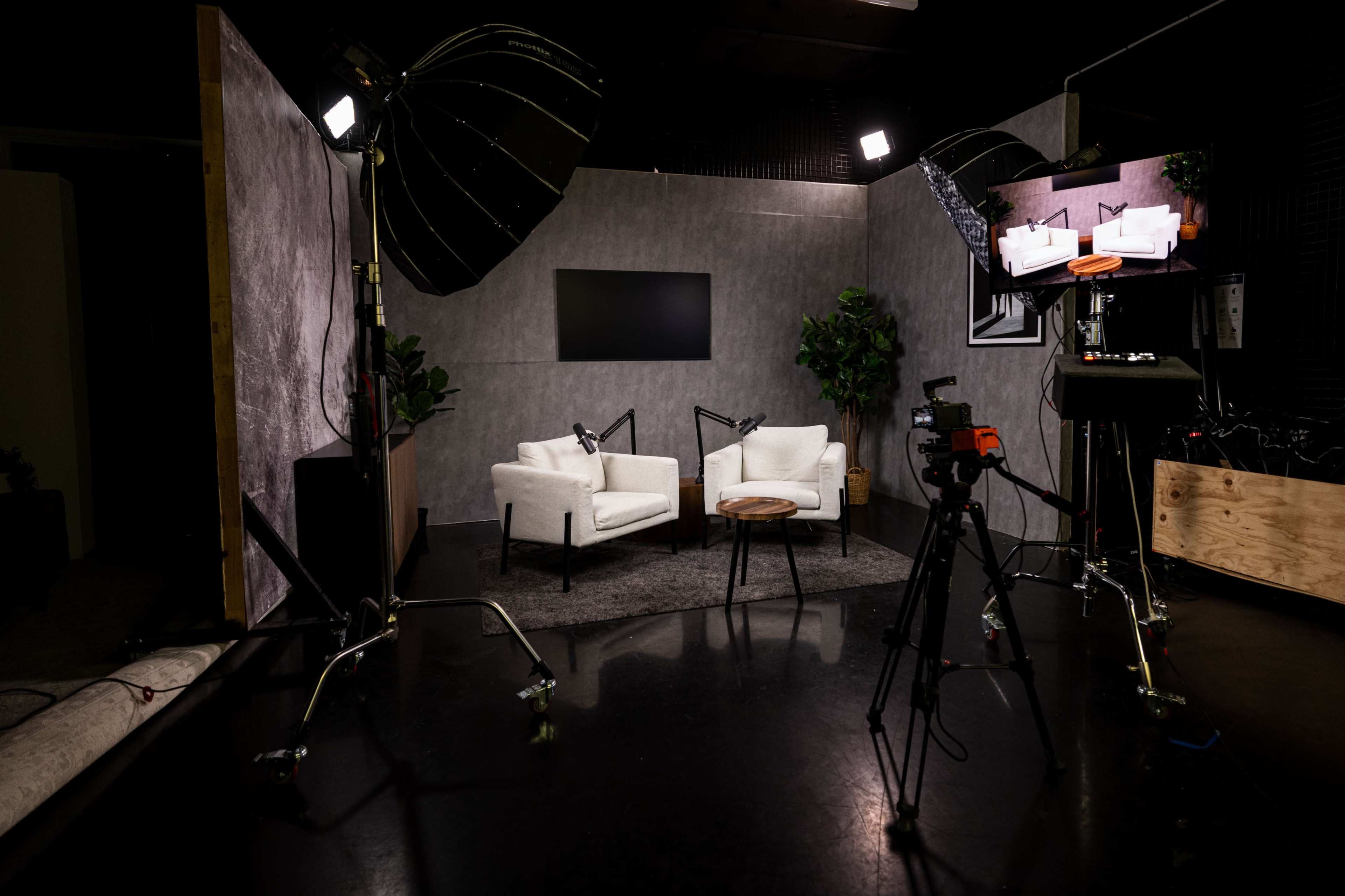 The image shows a video studio setup featuring two white chairs, a round wooden table, professional lighting, and camera equipment.