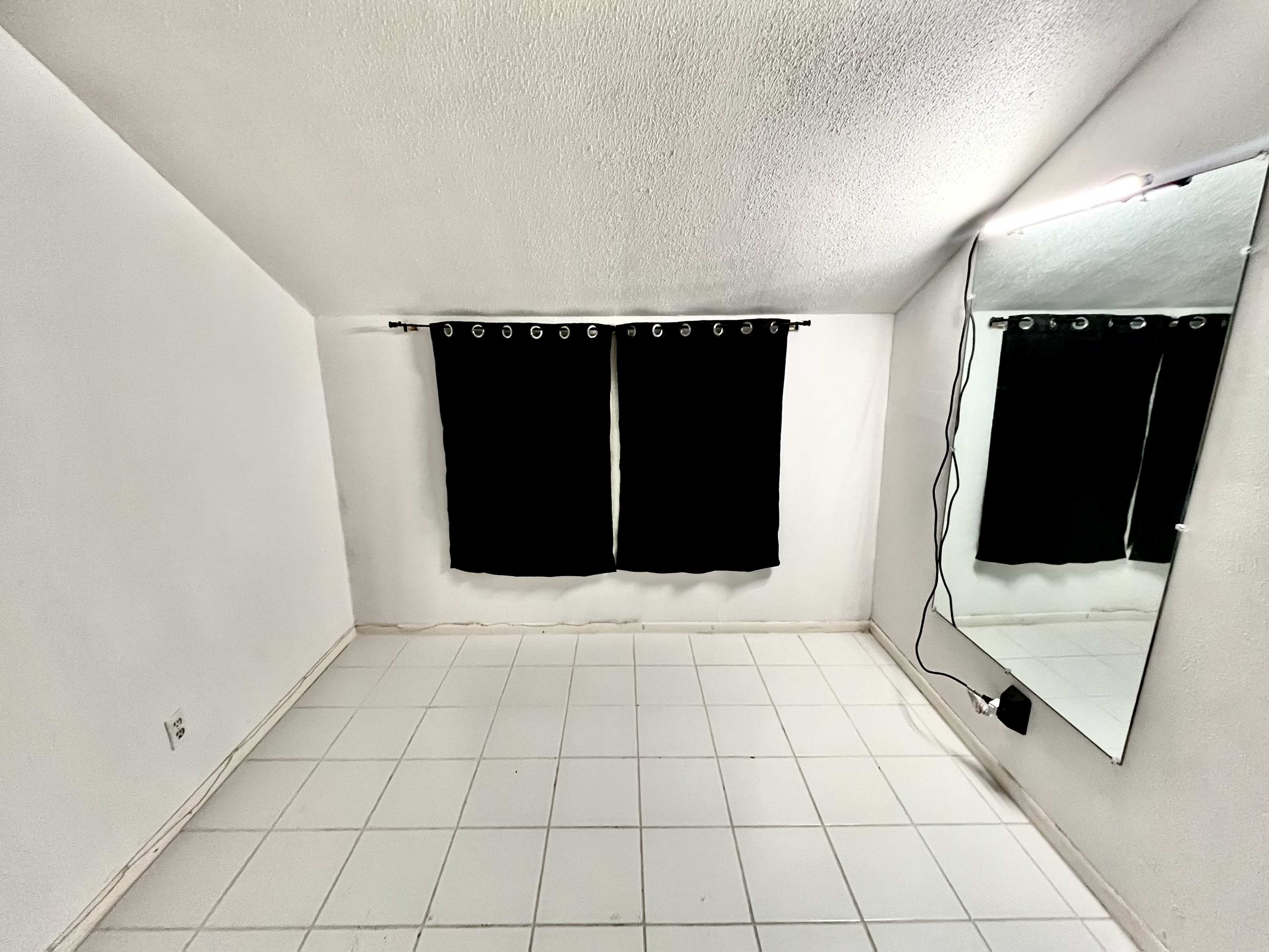 The image shows a sparsely furnished white room with two black curtains on a window and a large mirror mounted on the wall.