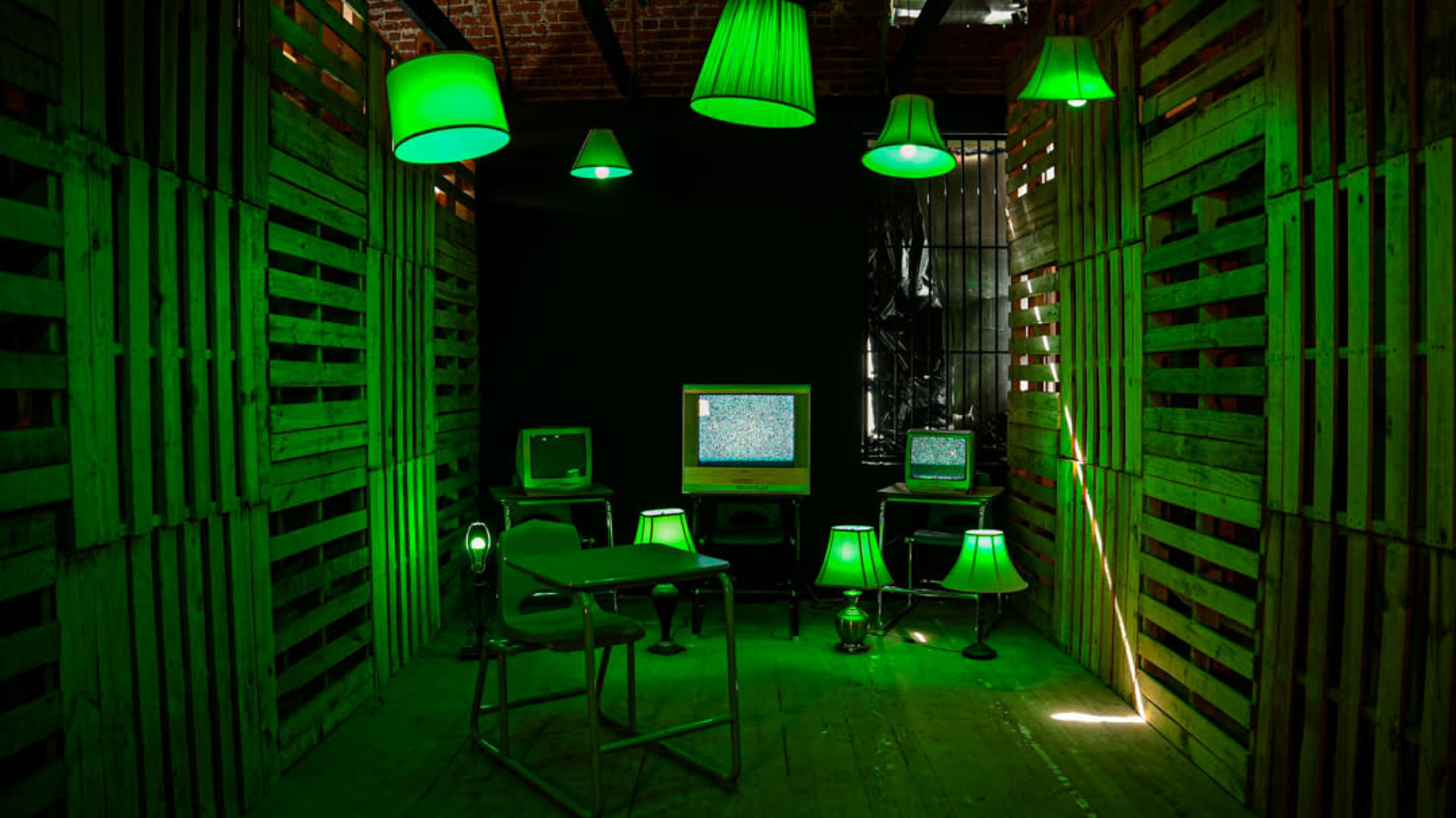 The scene features a dimly lit room with green lighting, showcasing multiple vintage televisions and lamps arranged around a small metal desk.