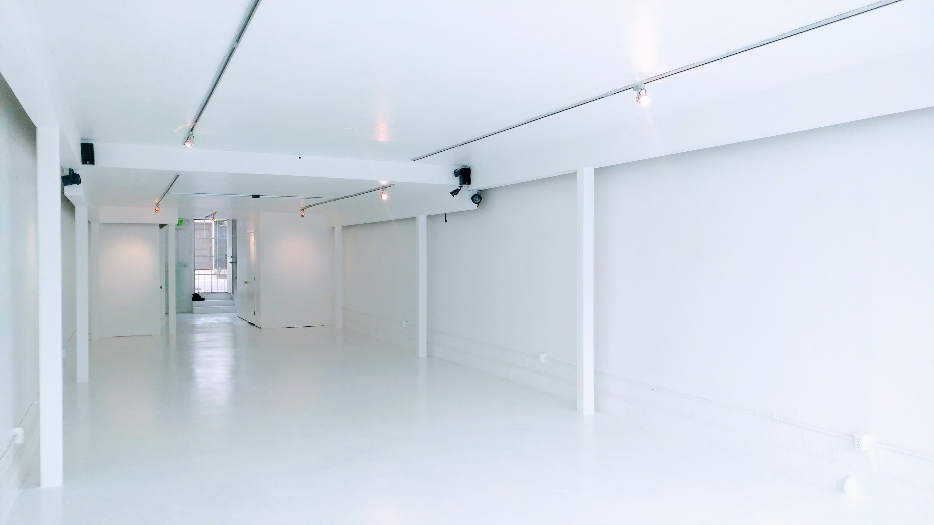 The image shows a spacious, minimalist room with white walls and a polished white floor, illuminated by overhead track lighting.