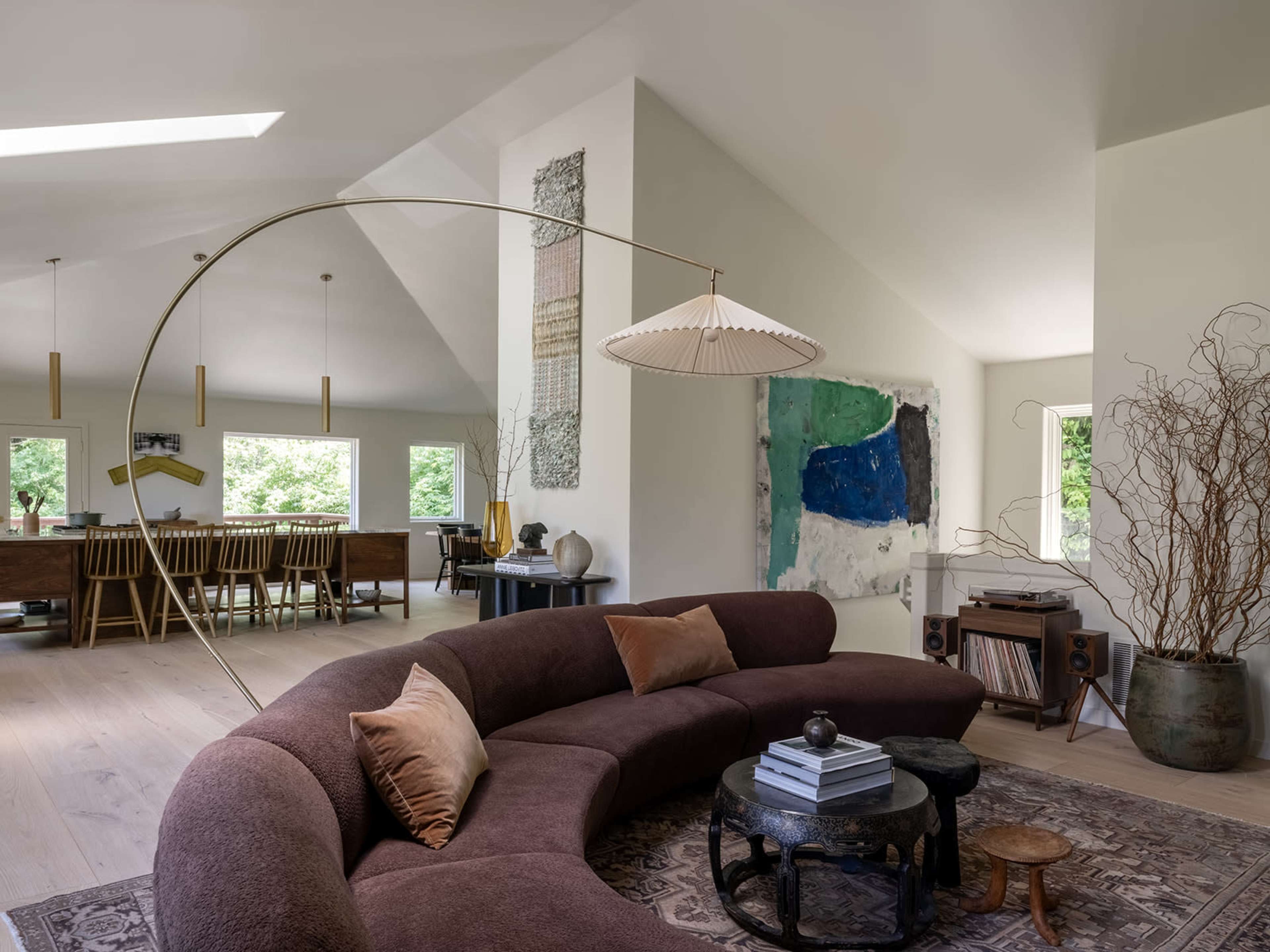 A modern living room features a curved brown sofa, a round coffee table, and a large abstract painting on the wall, with dining furniture visible in the background.