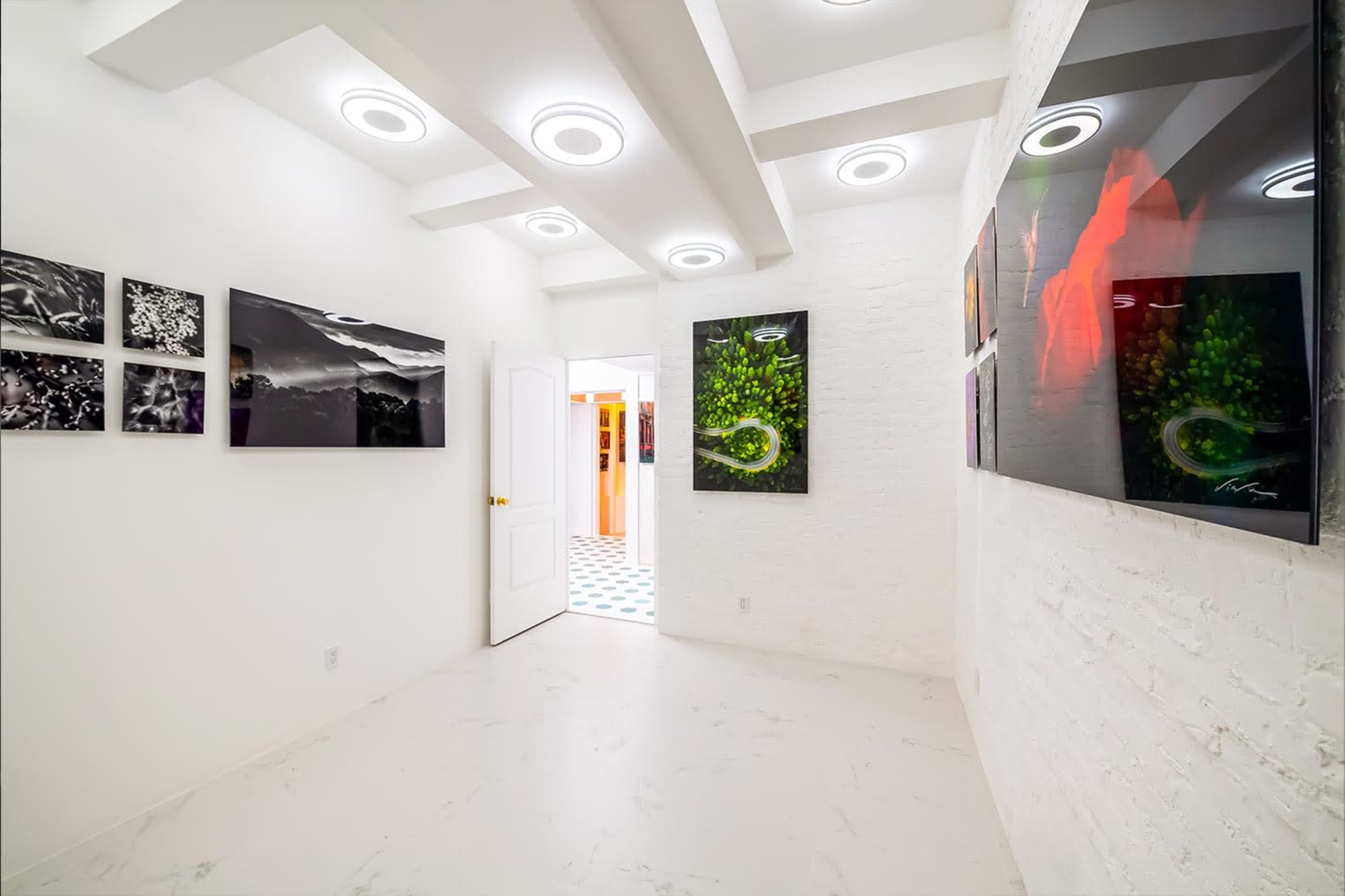 The image shows a white-walled gallery space featuring black and white photographs on one side and vibrant artwork displayed on the opposite wall, with a doorway leading to another room.