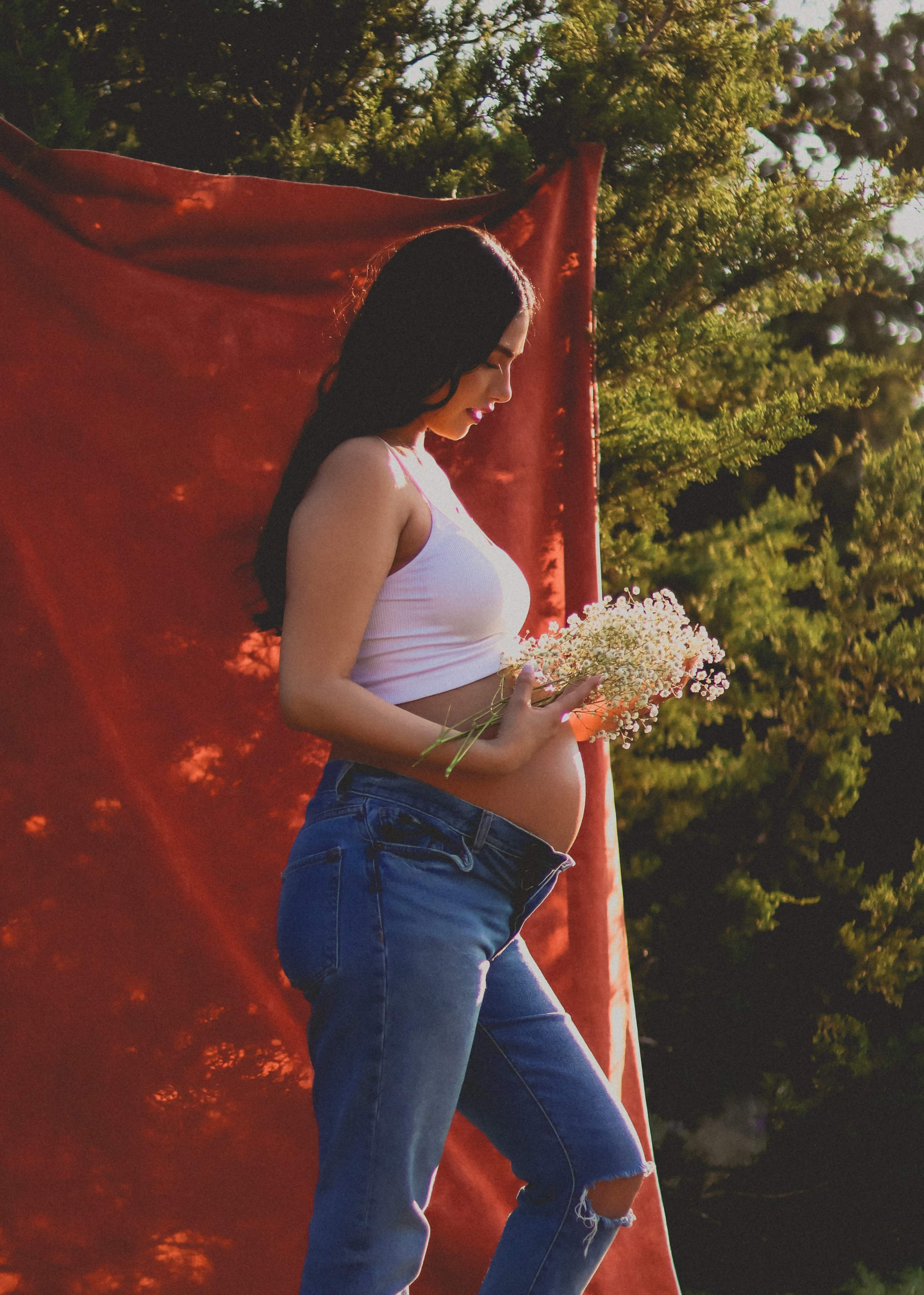 A pregnant woman wearing a white top and jeans holds a bouquet of flowers while standing in front of a red backdrop.