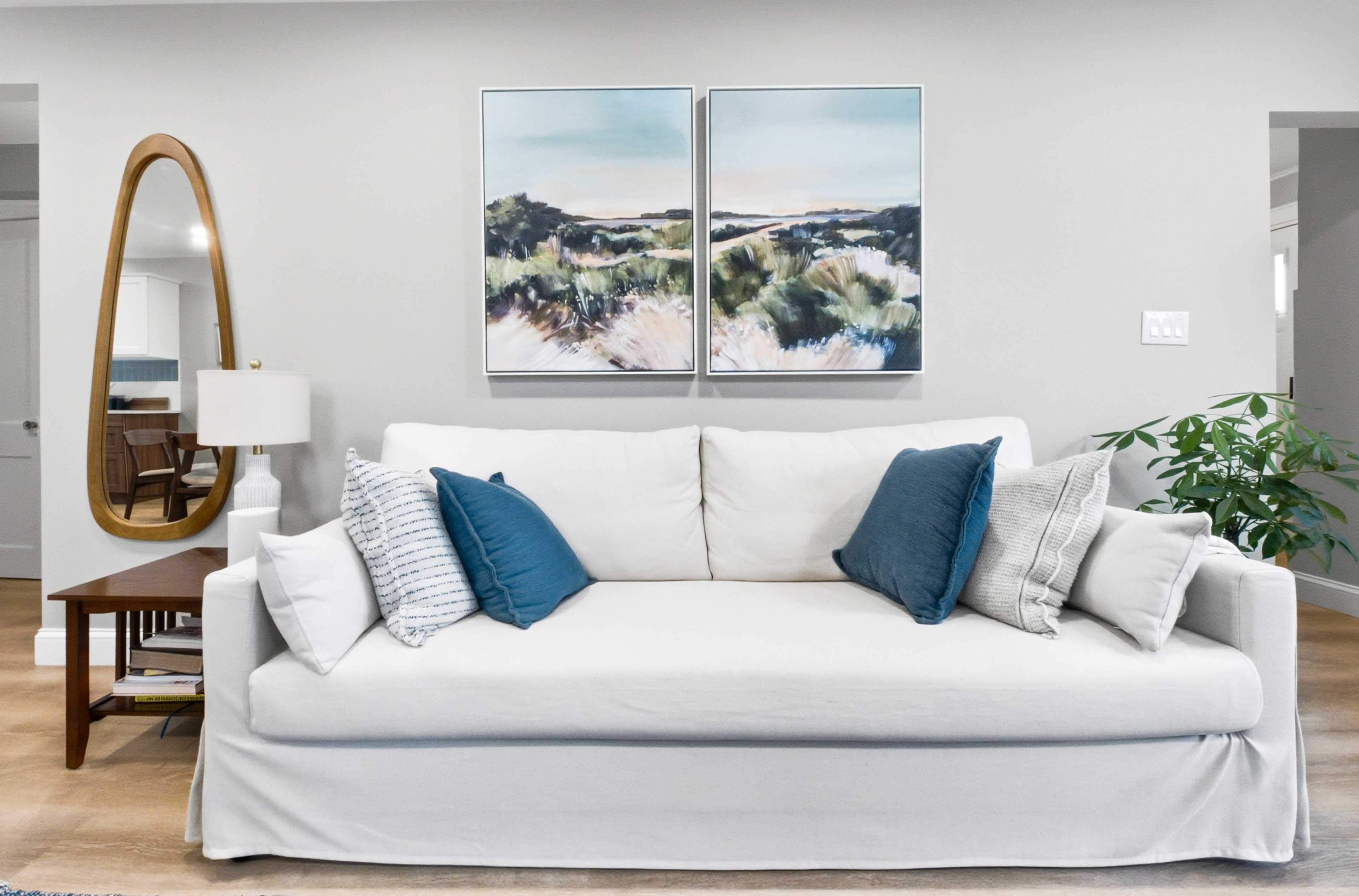 A white sofa with blue and gray pillows is positioned in a living room with a light-colored wall, a decorative mirror, a side table, and a plant.