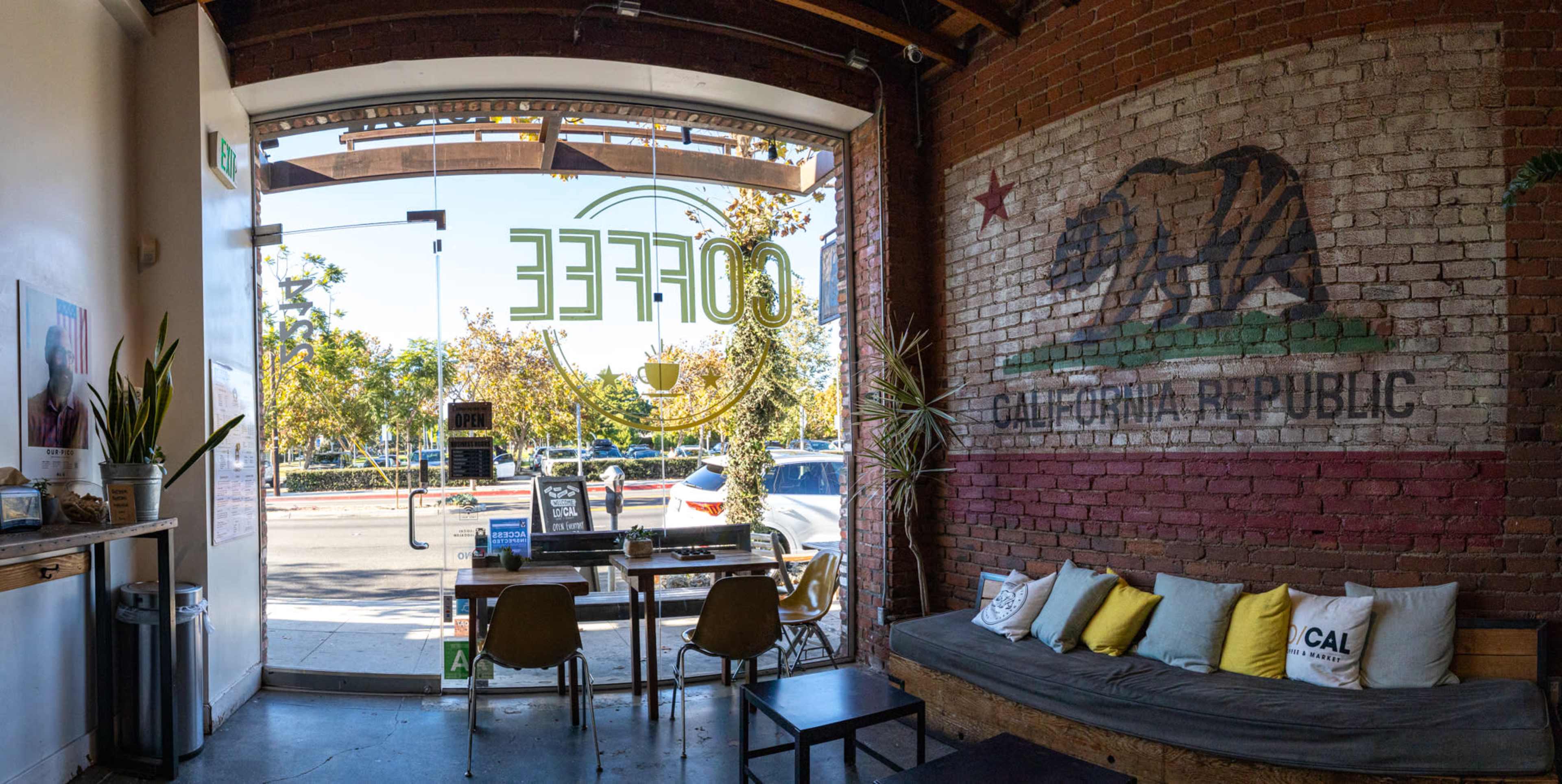 A coffee shop interior featuring a large mural of a bear and the words "California Republic" on a brick wall, with tables and chairs arranged near a glass entrance.