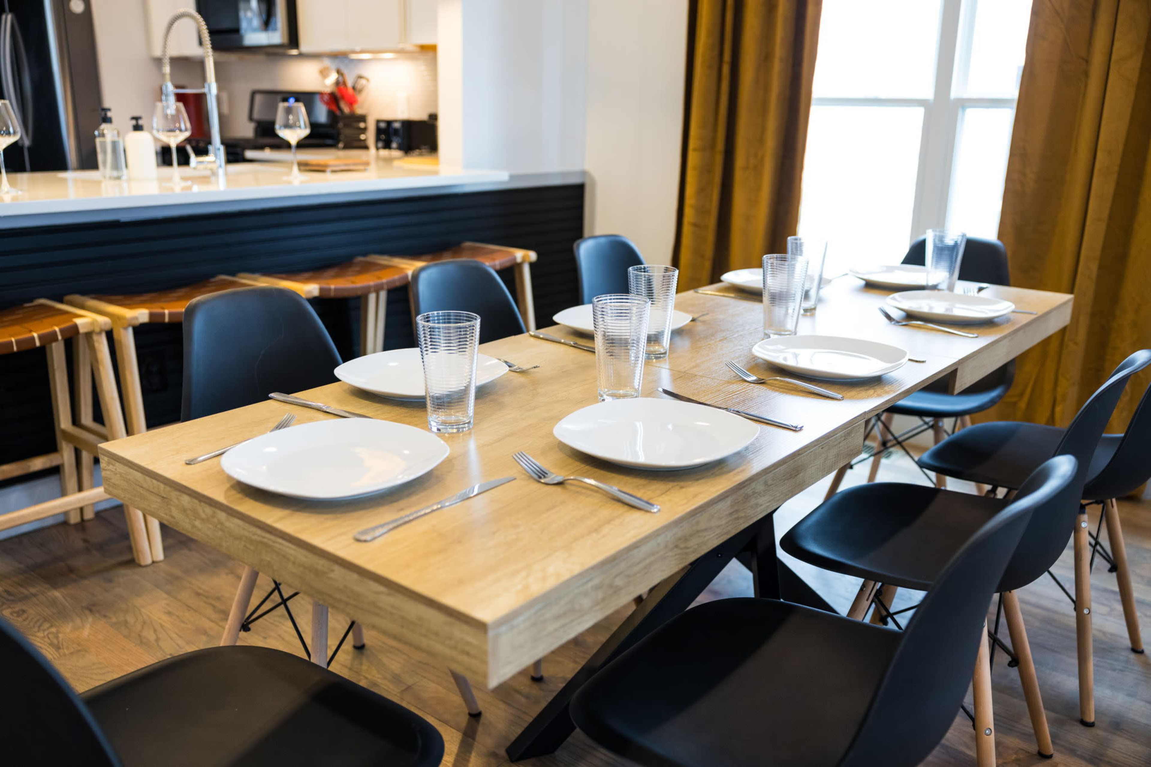 A wooden dining table is set with plates and glasses in a well-lit room with a kitchen visible in the background.