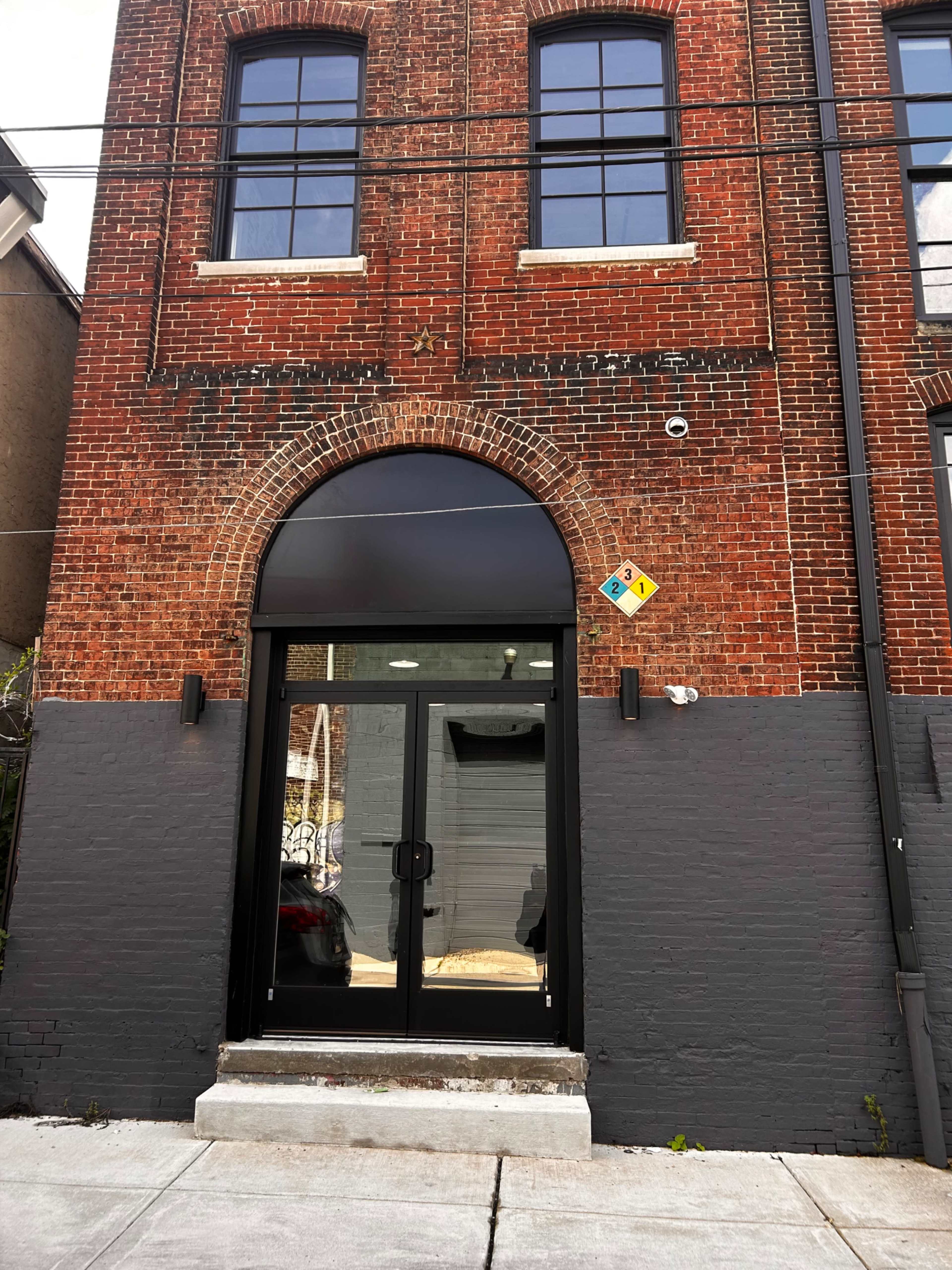 The image shows a brick building featuring large arched windows and a modern glass entrance door framed in black.